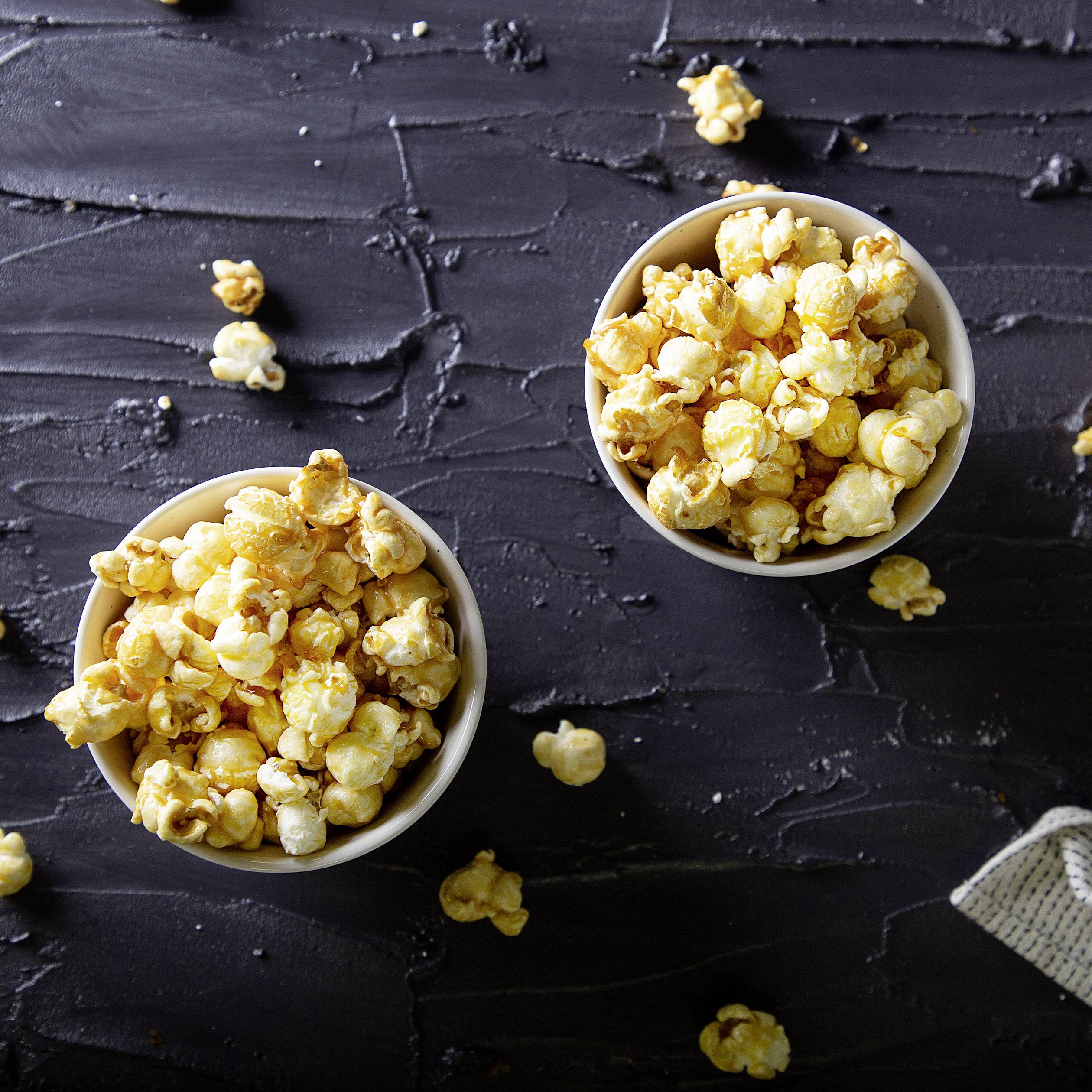 Dve misky s čerstvo pripraveným popcornom na tmavom povrchu, okolo ktorých sú rozhádzané jednotlivé kúsky popcornu.