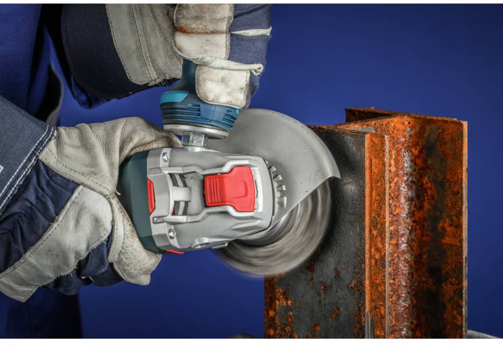 Osoba v rukaviciach brúsi hrdzu z kovového nosníka uhlovou brúskou. Pozadie je modré.
