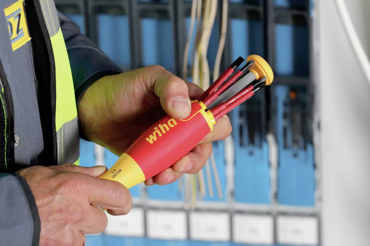 Osoba drží skrutkovač s viacerými nástavcami pred otvorenou elektrickou rozvodnou skriňou. Na skrutkovači je viditeľná značka 'Wiha'.