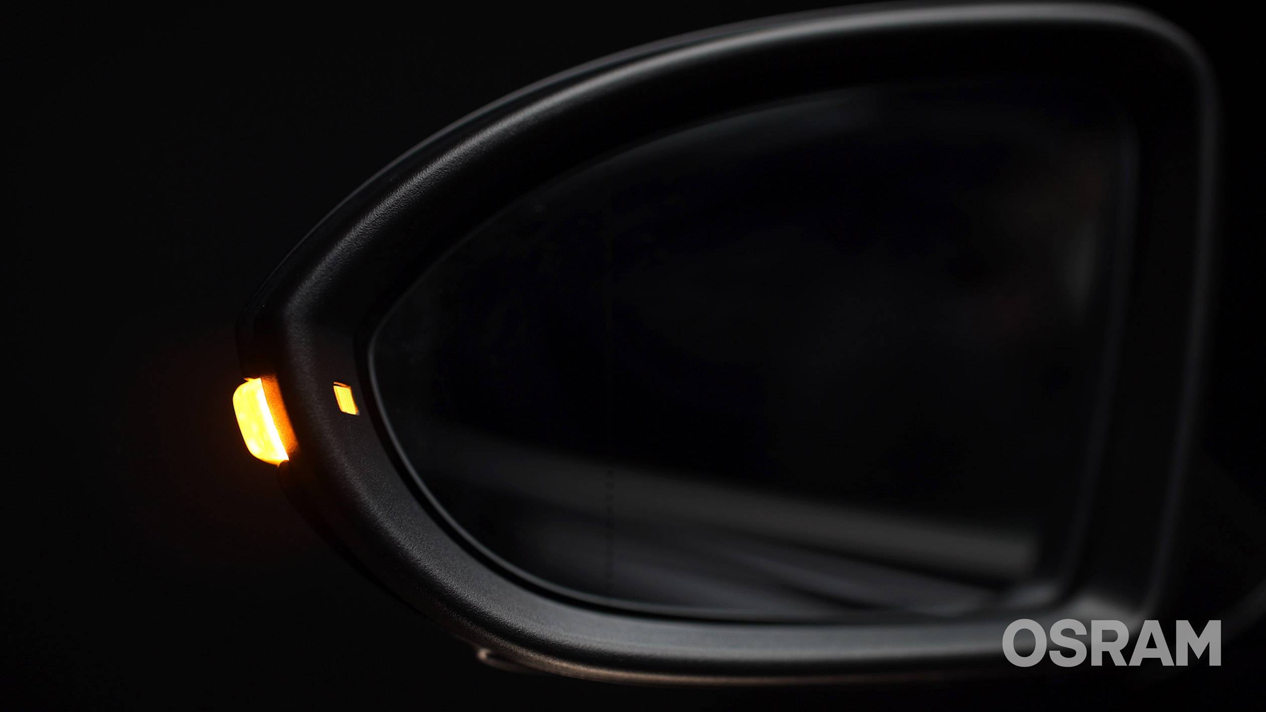 Pohľad z bočnej strany spätného zrkadla automobilu so svietiacim výstražným signálom na tmavom pozadí. Logo OSRAM sa nachádza v pravom dolnom rohu obrázka.