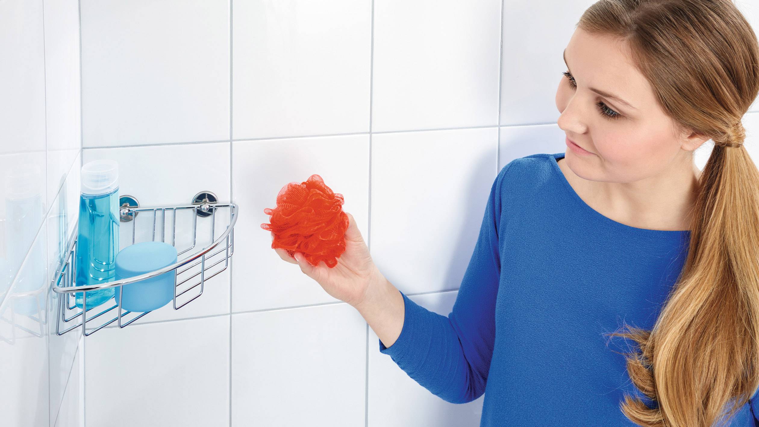 Žena v modrom oblečení drží v ruke oranžovú sprchový hubku, stojí pred sprchovým regálom s modrou fľašou na sprchový gél a dózou.