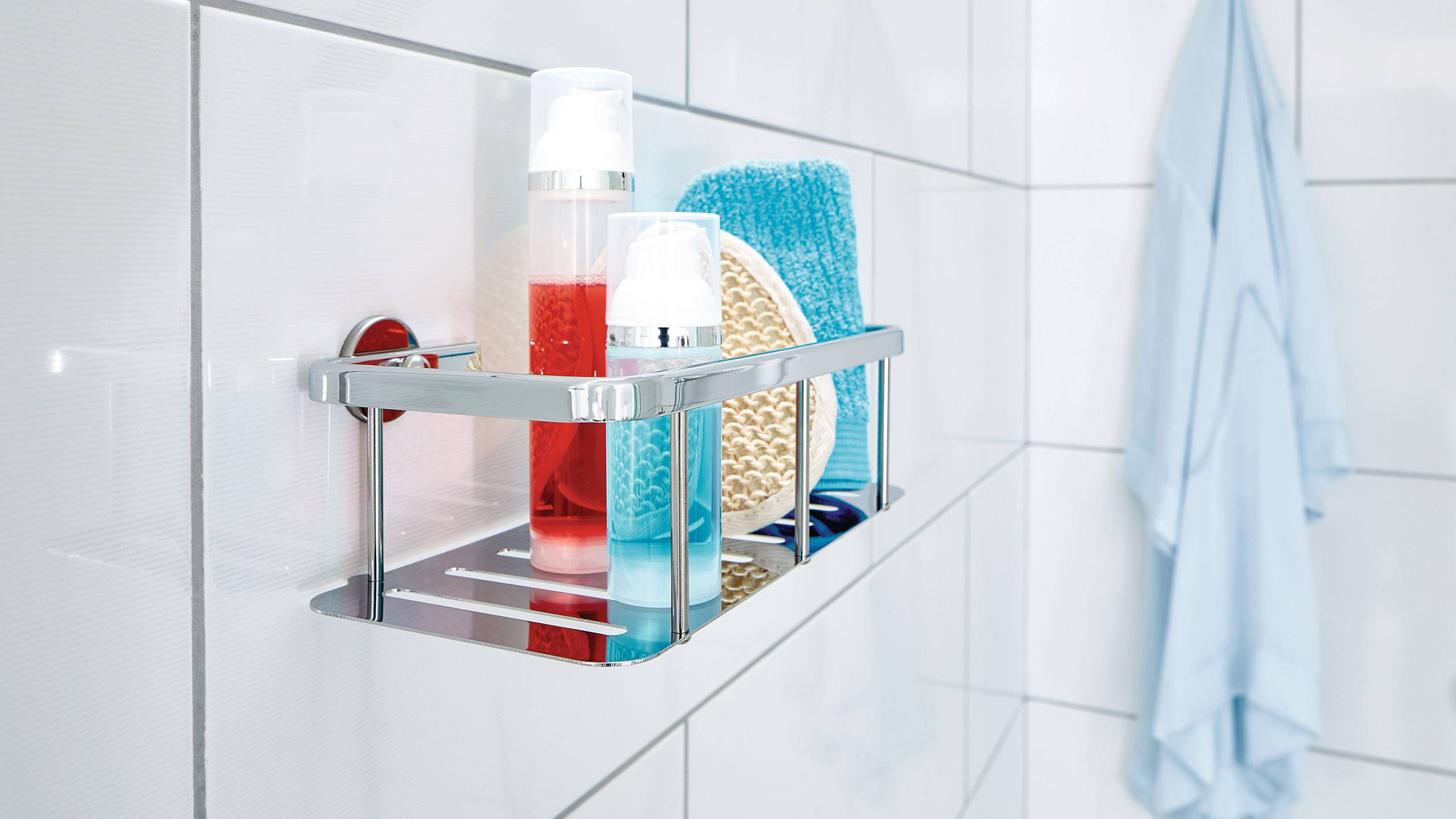 Kúpeľňová polica so sprchovým gélom, špongiou a ďalšími hygienickými produktmi na bielej obkladačkovej stene. Vedľa visí modré uterák.