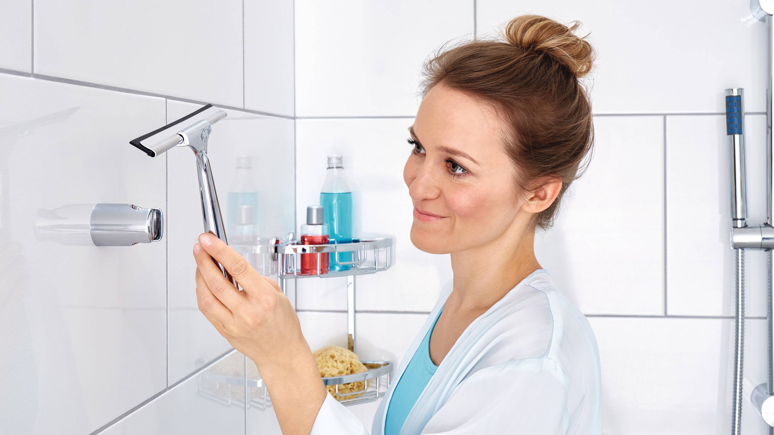 Žena čistí dlaždice v kúpeľni stierkou. Vedľa nej sú hygienické produkty uložené na poličke v sprche.