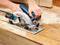 Osoba používa Boschovu okružnú pílu na drevenom pracovnom stole, ktorá prechádza kusom dreva, pričom okolo sú roztrúsené piliny.