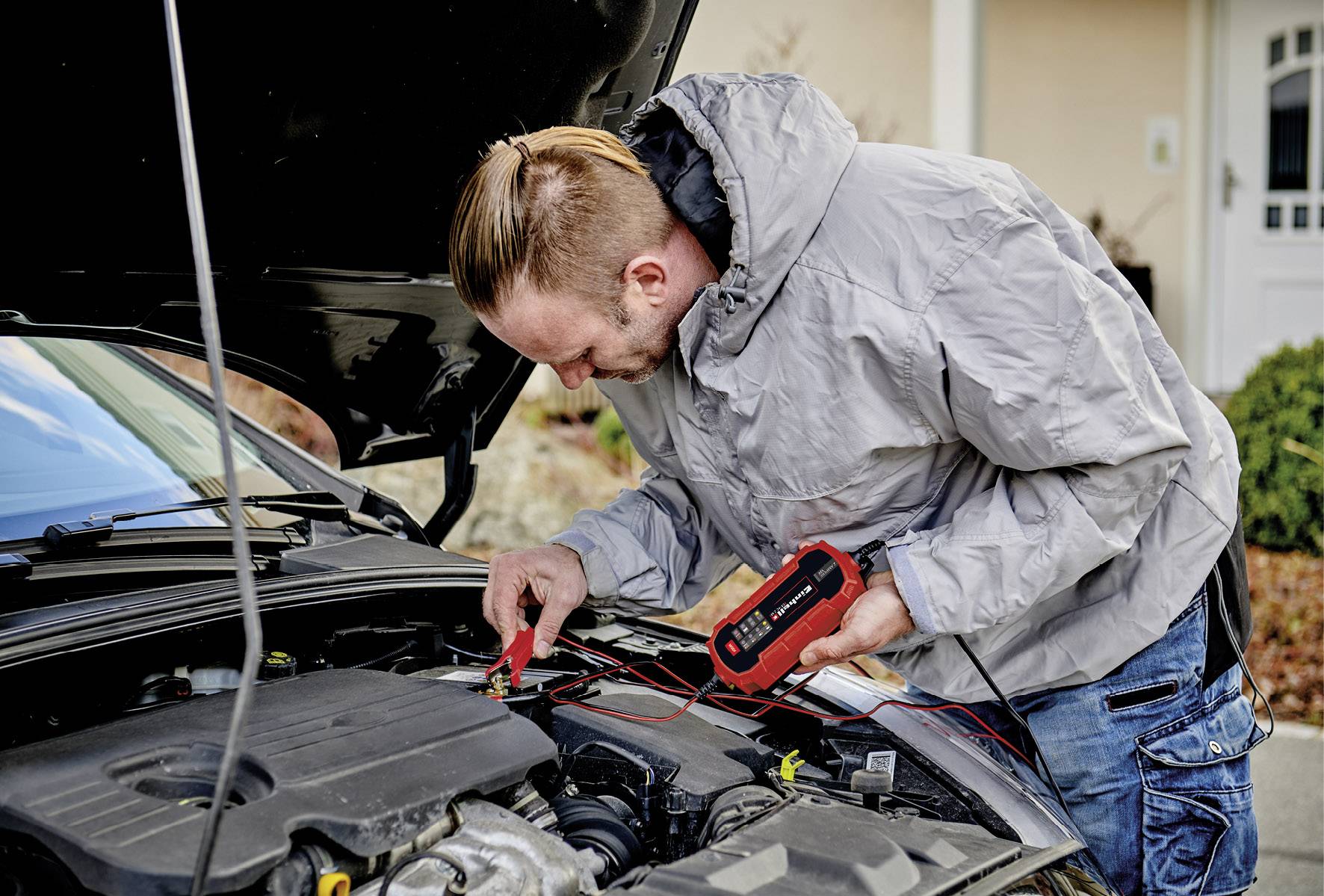 'Muž testuje autobatériu elektrickým testovacím zariadením pri otvorenej kapote pred domom'