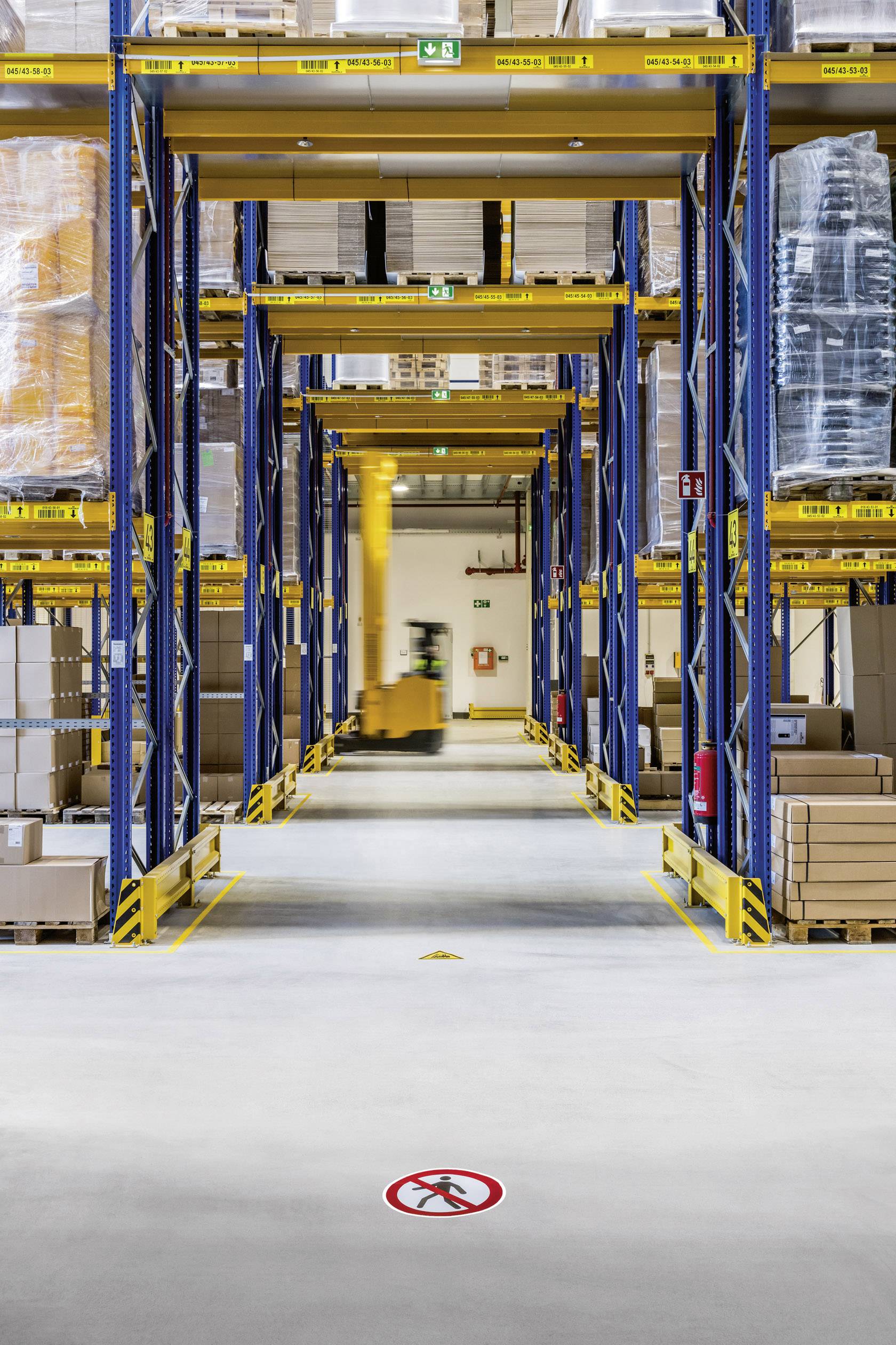 Široká chodba v sklade s vysokými regálmi plnými krabíc. Vo vzdialenosti sa pohybuje vysokozdvižný vozík.