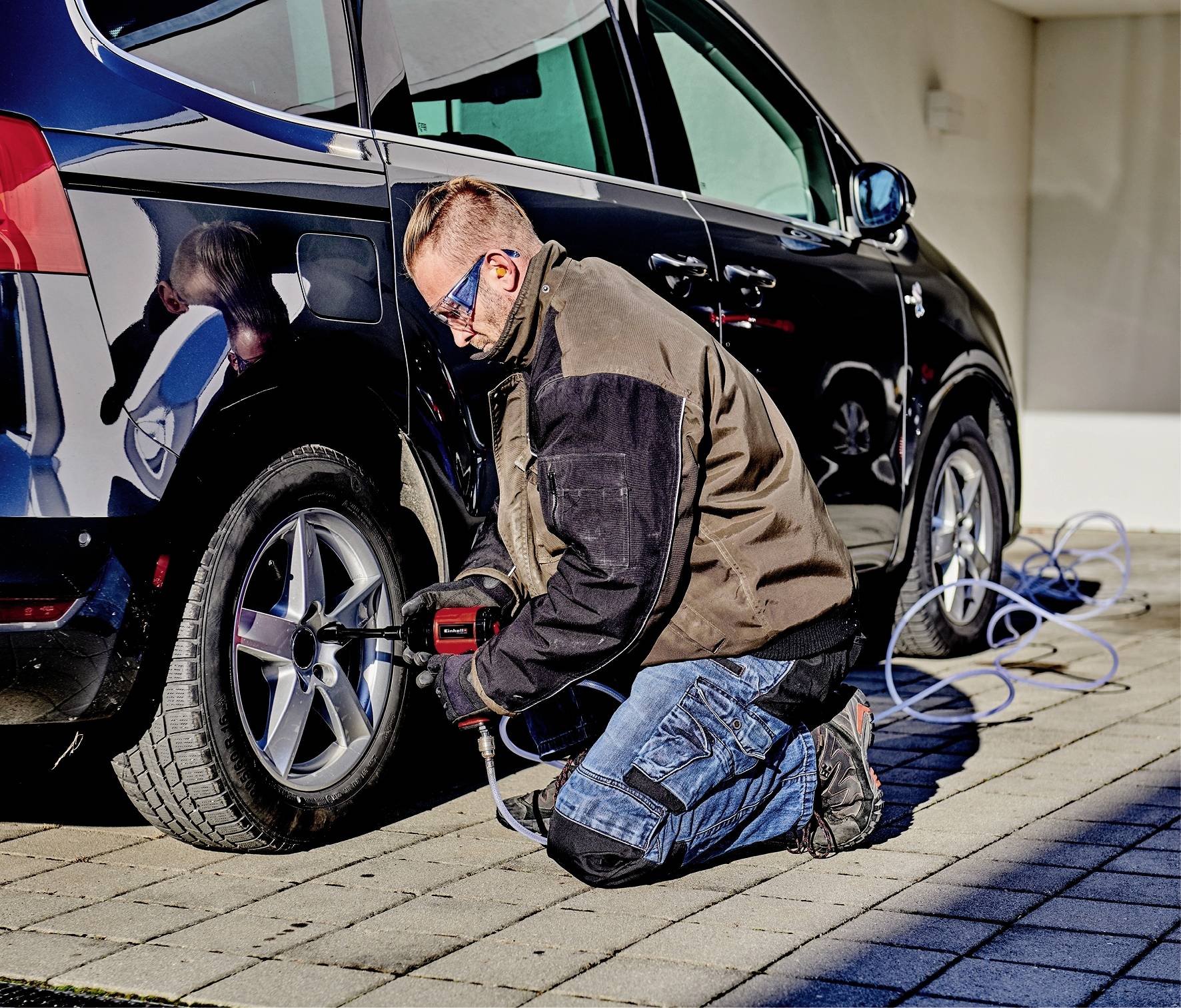 Osoba kľačí na zemi a používa nástroj na kontrolu alebo opravu pneumatík čierneho automobilu.