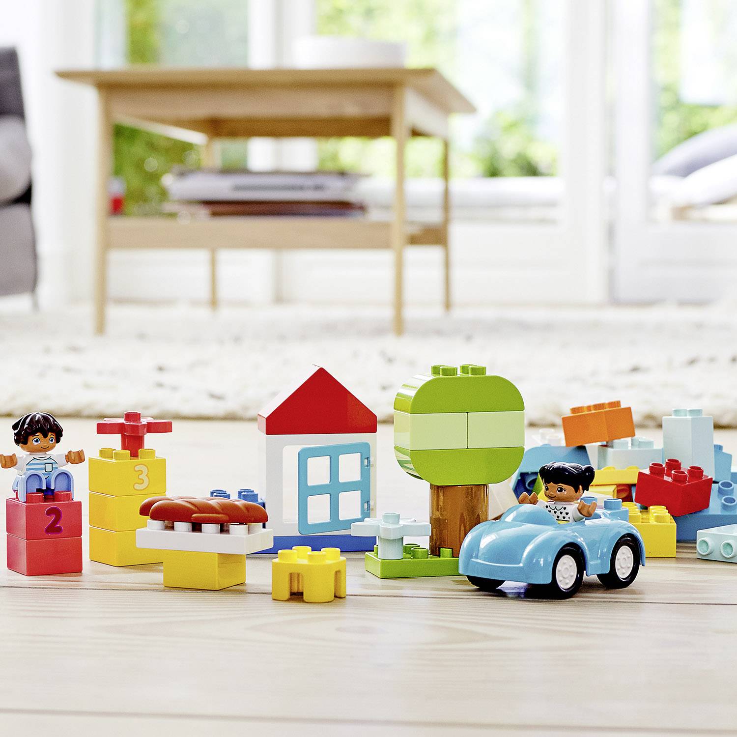 Farebná LEGO súprava na podlahe s figúrkami, autom, stromami a domom. V pozadí stôl a okno.