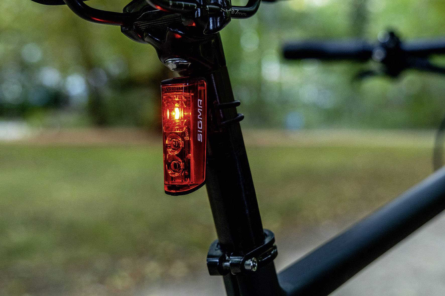Bicykel s rozsvietením červeným zadným svetlom, namontovaným na sedlovej trubke. Rozmazané pozadie s drevinami a trávou.