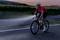 Cyklista v červenom drese a s helmou jazdí po ceste za súmraku. Svetlo jeho bicykla osvetľuje cestu pred ním. V pozadí je vidieť rad stromov.