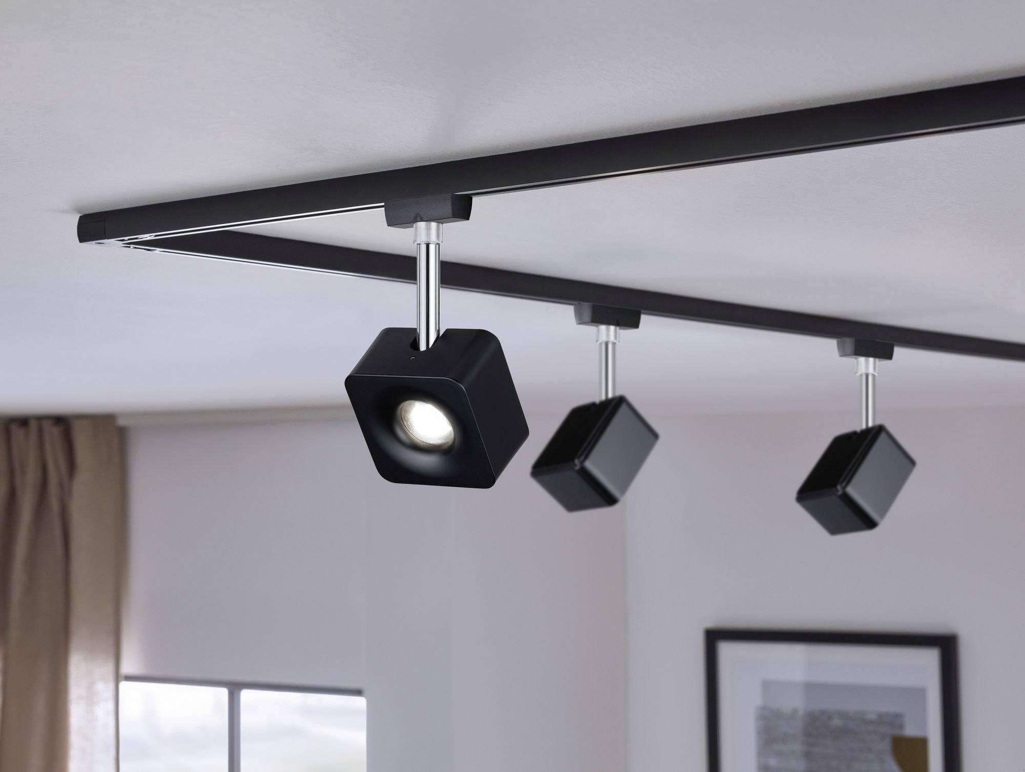 Tri čierne stropné reflektory na lište osvetľujú priestor s modernými akcentmi, vrátane zarámovaného obrazu na stene.