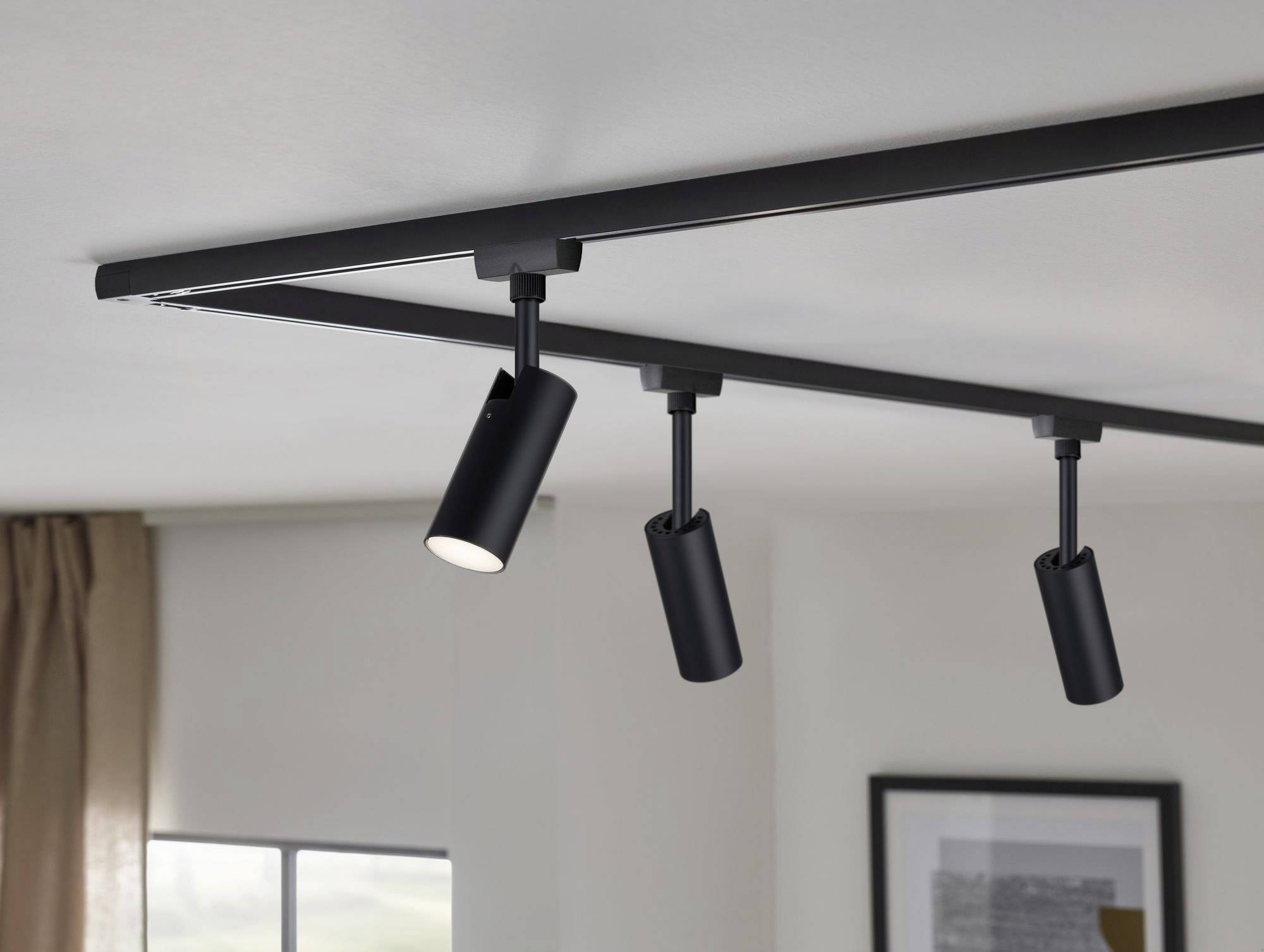 Čierne koľajnicové svietidlo s troma nastaviteľnými bodovými svetlami na strope miestnosti, pred oknom so závesom a rámom obrazu.