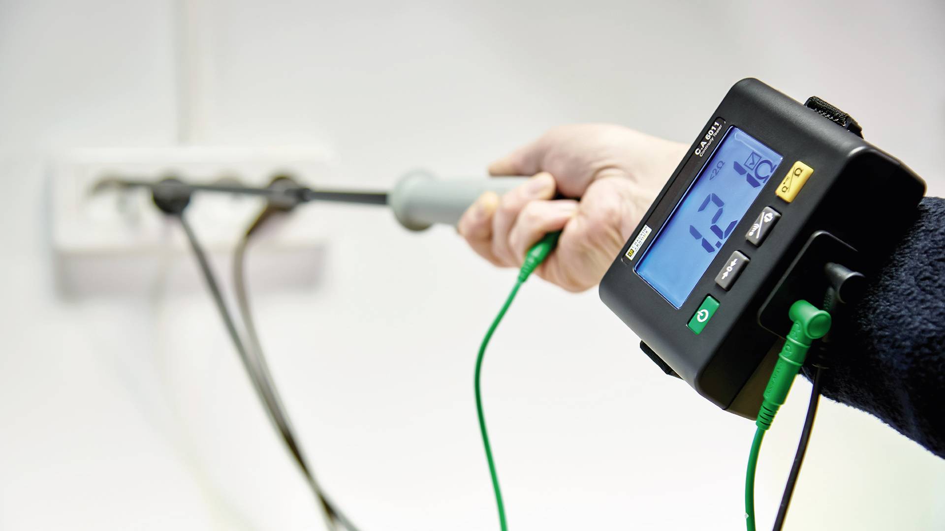 Jedna ruka drží merač napätia pripojený do elektrickej zásuvky. Displej zobrazuje '123 V'.