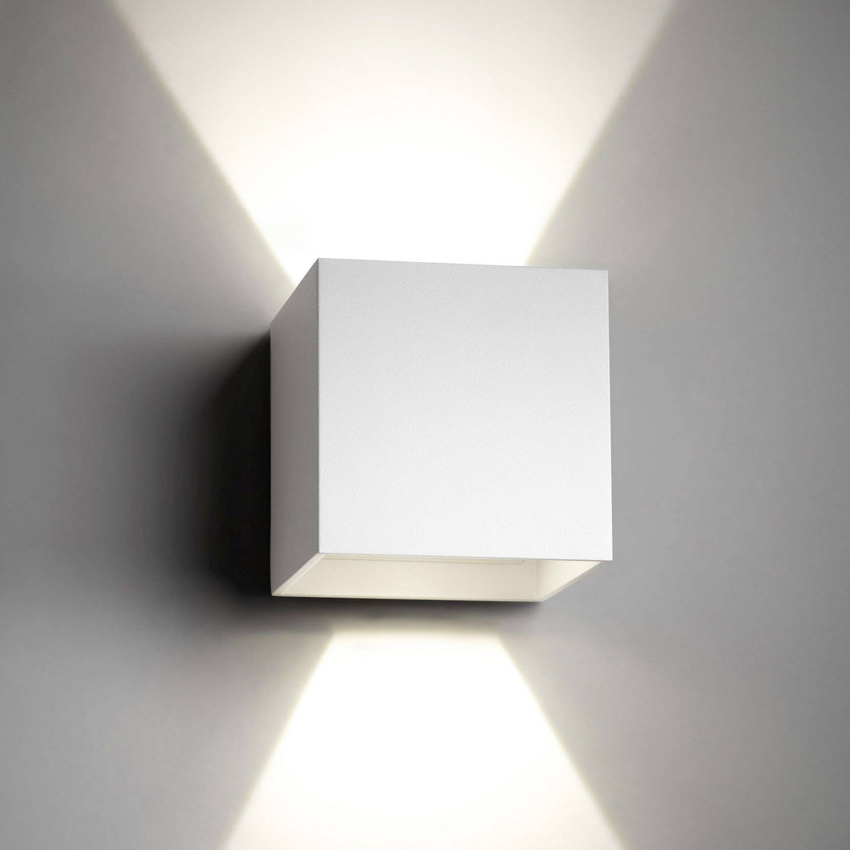Stenová lampa v tvare kocky z bieleho kovu, vyžaruje teplé svetlo smerom nahor a nadol. Minimalistický dizajn na sivej stene.
