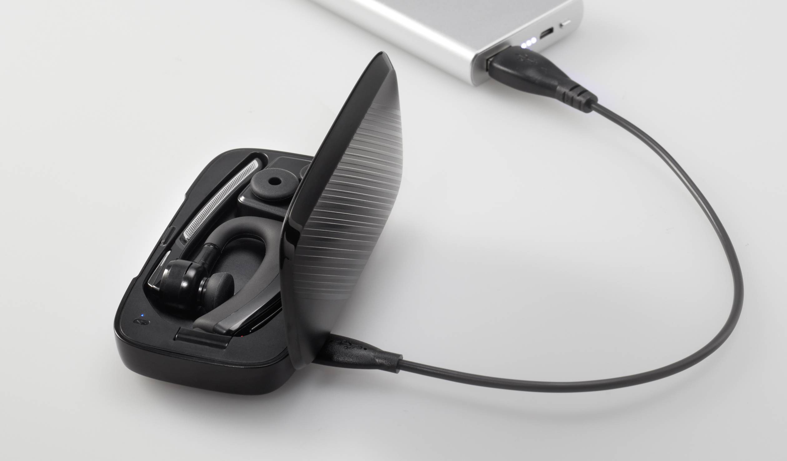 Bluetooth headset leží v otvorenom nabíjacom puzdre, ktoré je prepojené USB káblom s powerbankou.