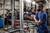 Muž vŕta elektrickým vŕtacím strojom do kovového rámu v dielni. V pozadí pracuje ďalšia osoba.