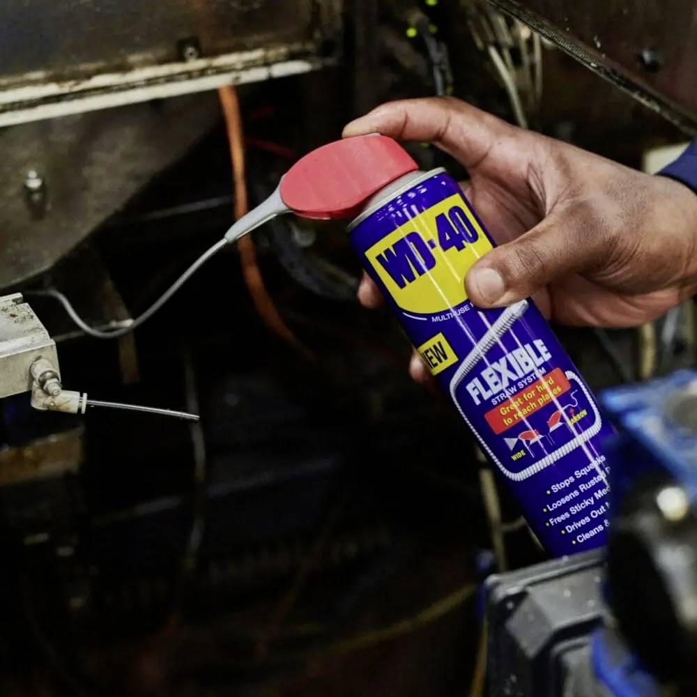 Jedna ruka drží sprejovú nádobu WD-40 FLEXIBLE s ohybnou sprejovacou trubicou. Osoba práve nanáša produkt na strojovú súčiastku.