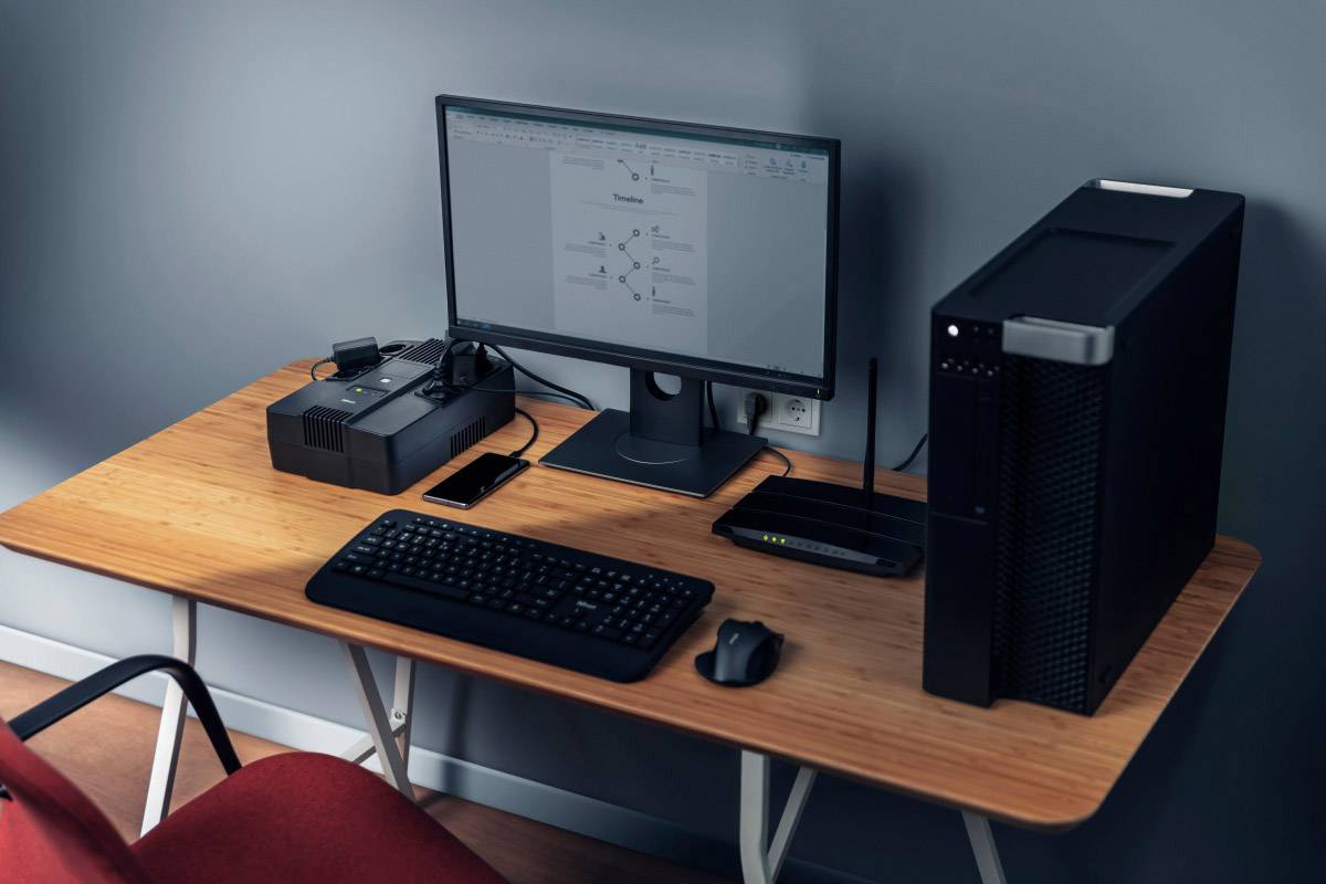 Písací stôl s počítačom, monitorom, klávesnicou, myšou, smartfónom a smerovačom. Monitor zobrazuje dokument.