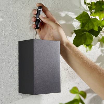 Ruka montuje čiernu lampu na bielej stene pomocou skrutkovača; v pozadí zelené rastliny.