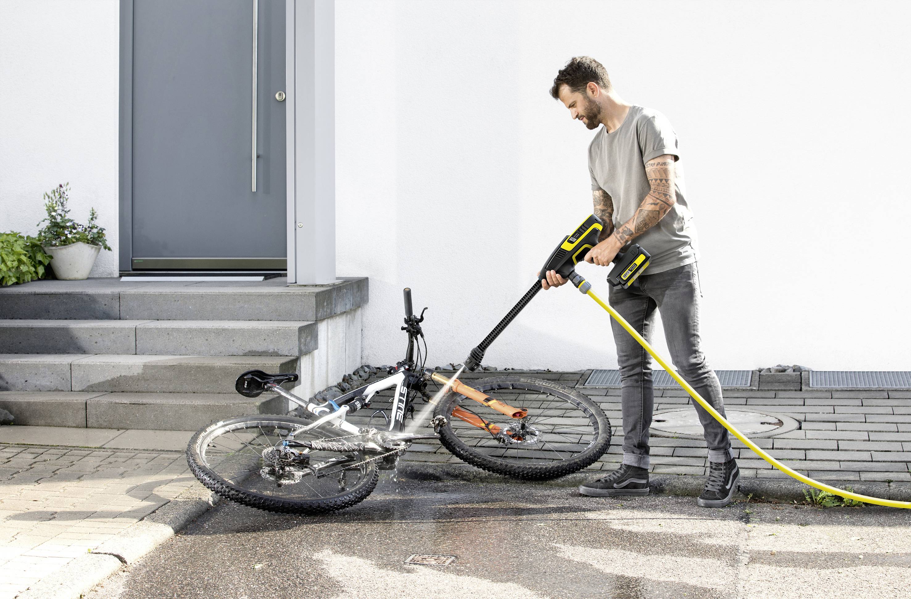 Muž čistí bicykel vysokotlakovým čističom na mokrej ceste vedľa schodiska.