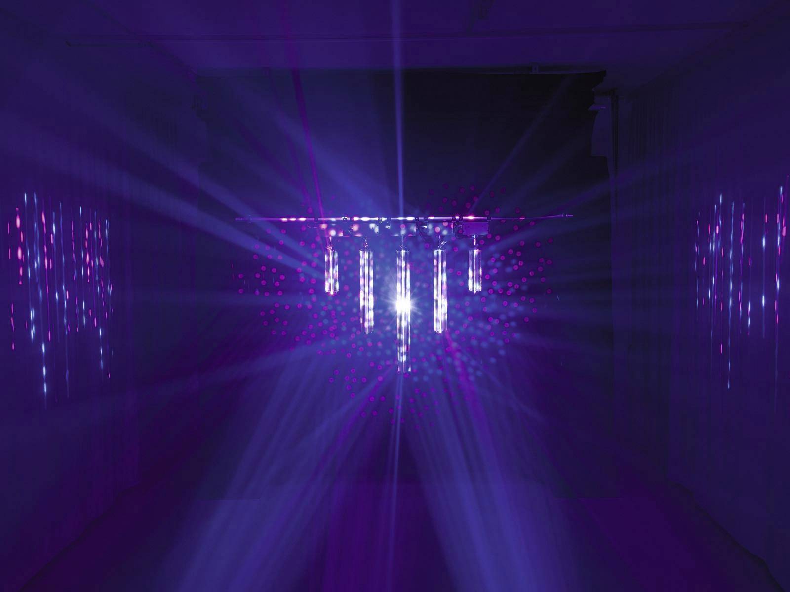 Niekoľko vertikálnych svetelných lúčov v modrej a ružovej farbe vyžaruje zo stredu tmavej miestnosti a vytvára trblietavý efekt.