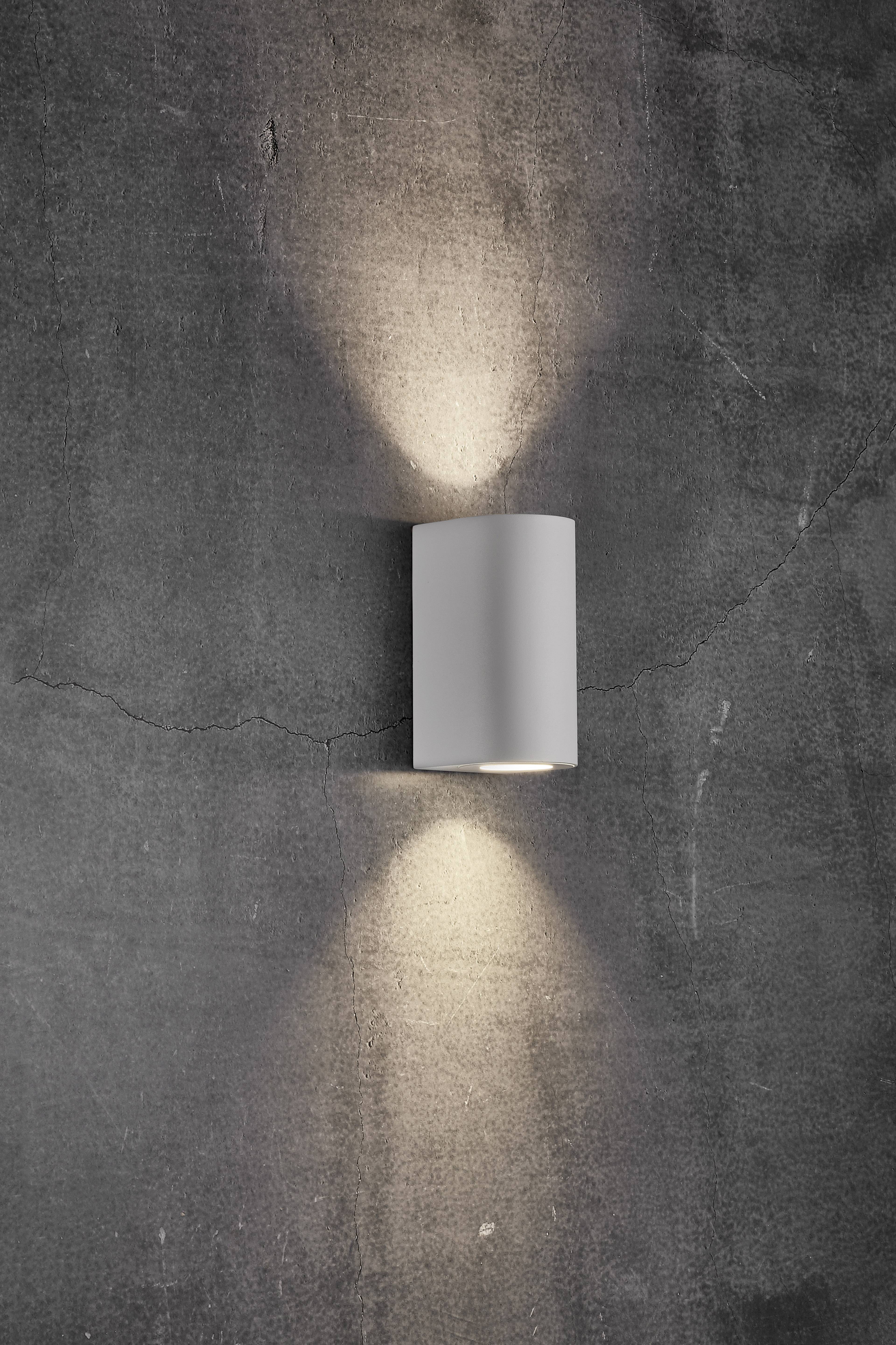 Valcová, na stenu namontovaná lampa v modernom dizajne, ktorá vyžaruje svetlo smerom nahor aj nadol na sivej betónovej stene.
