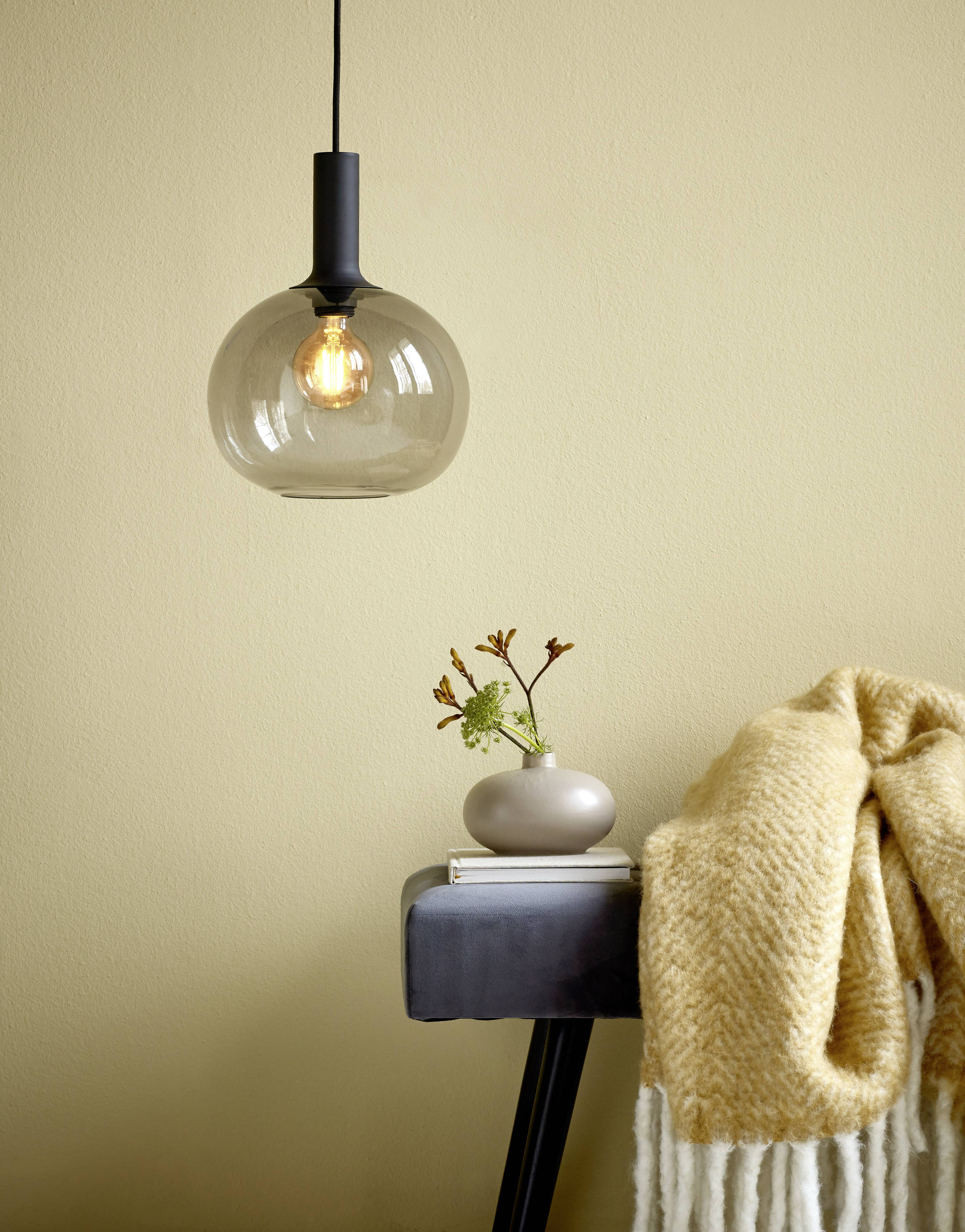 Závesná lampa z šedého skla nad stolom s vákou a kvetmi. Vedľa leží béžová deka. Jednoduchý, moderný interiér.