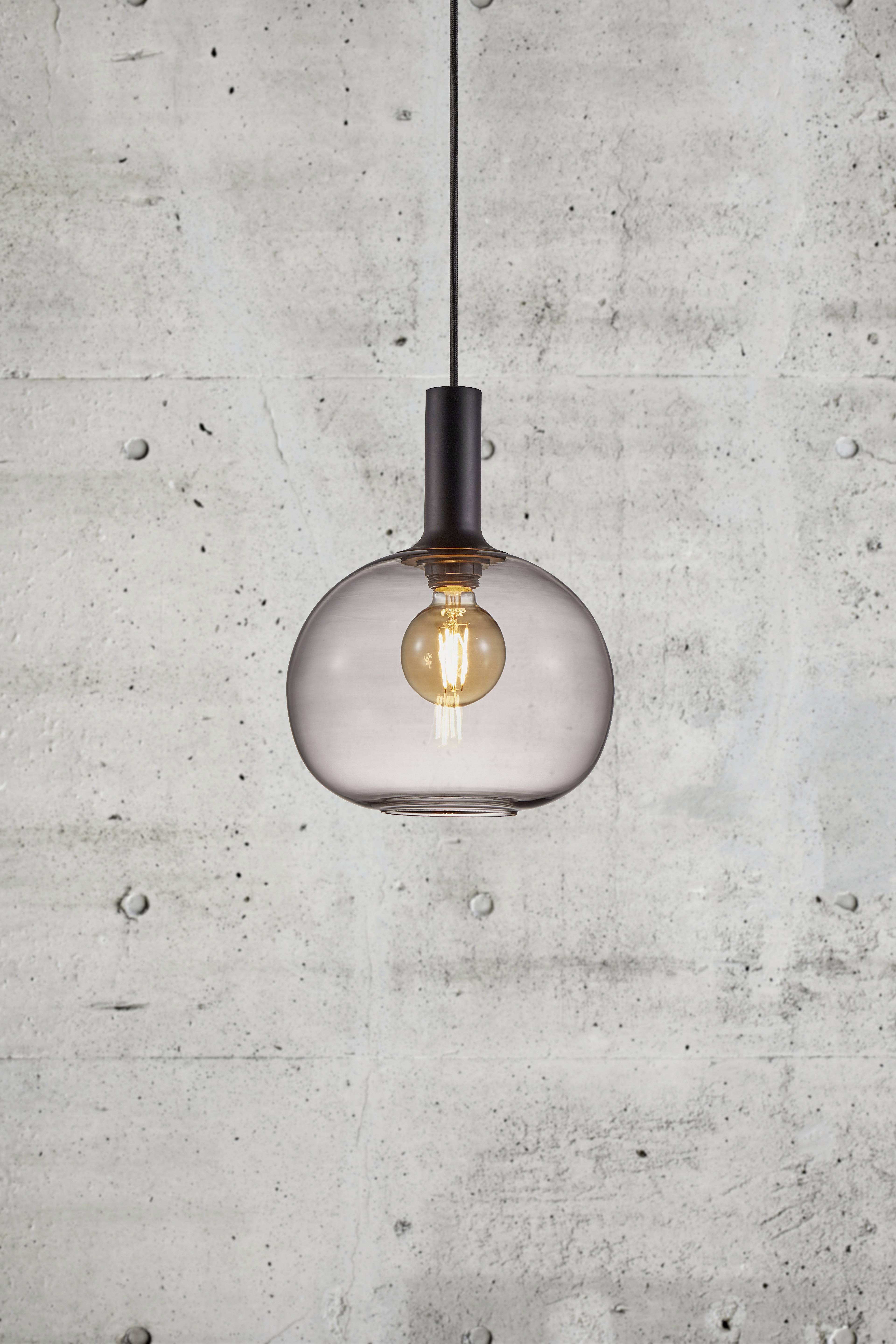 Guľovitá závesná lampa so viditeľným svetelným zdrojom visí pred betónovou stenou.