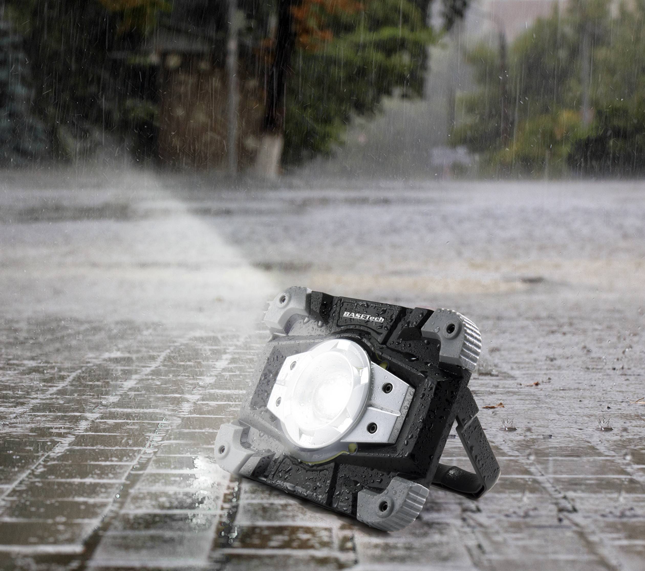 Odolná pracovná lampa stojí na mokrej dlažbe v daždi, vyžaruje jasné svetlo a osvecuje okolie.