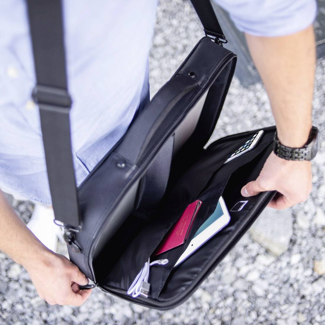 Osoba otvára čiernu tašku cez rameno s laptopom, smartfónom a poznámkovým blokom vo vnútri, na štrkovej ceste.