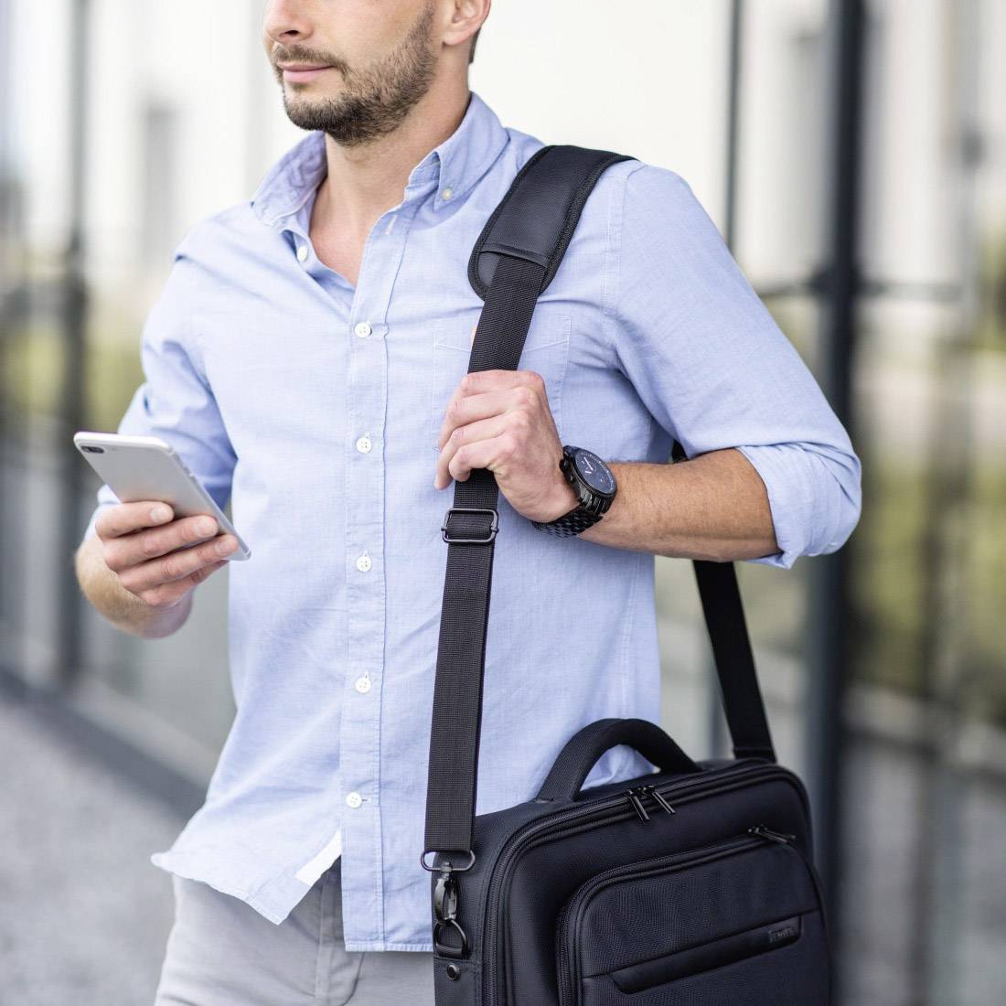 Muž s bradou a v zatvorenom svetlomodrom tričku stojí vonku, drží smartfón a má prevesenú tašku cez plece.