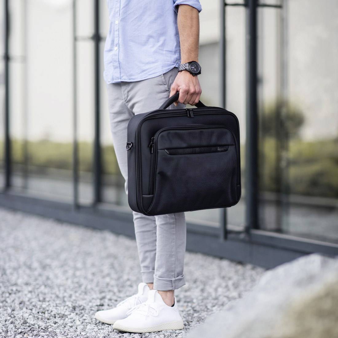 Osoba nesie čiernu tašku cez rameno a stojí na štrku pred modernou presklenou budovou.