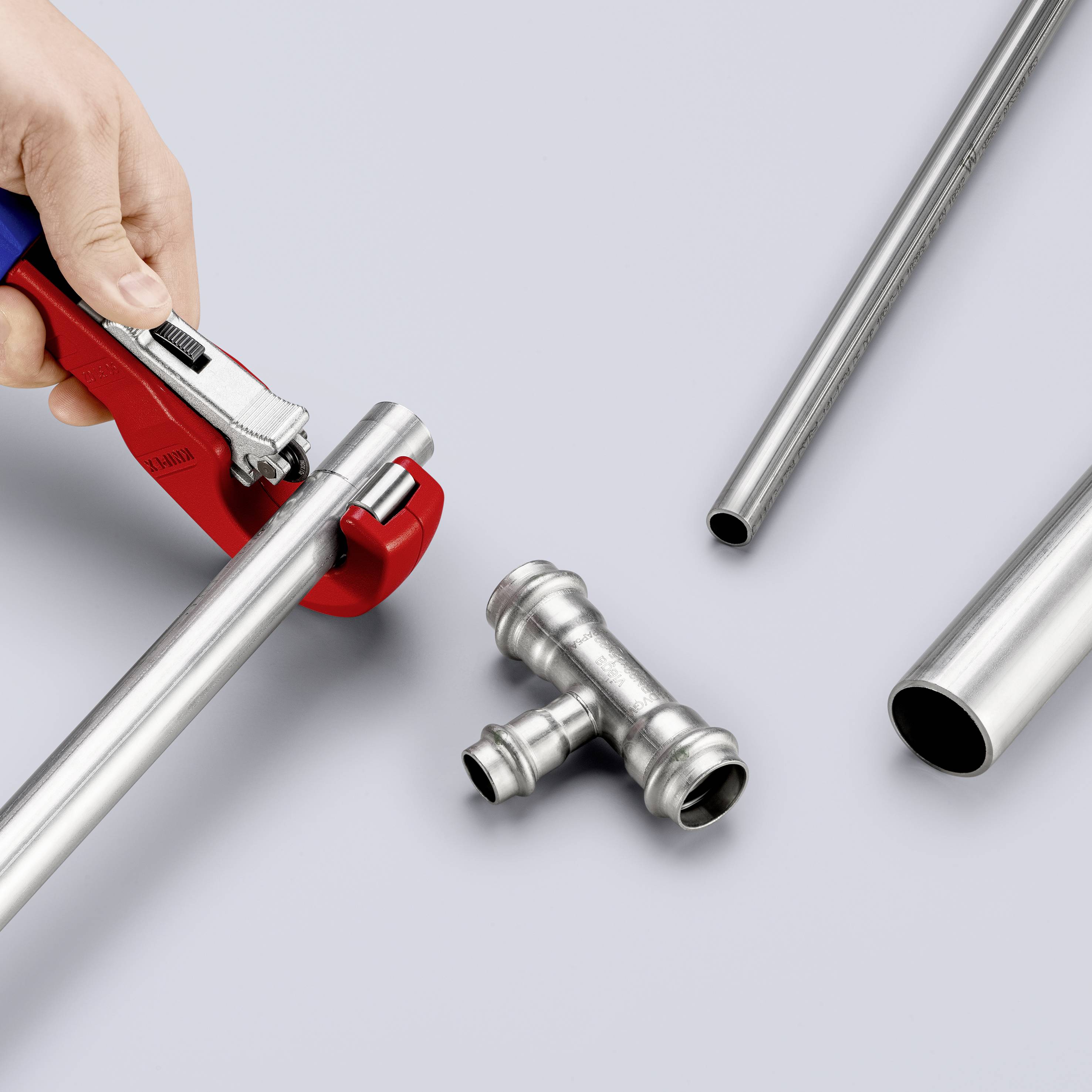 Osoba strihá kovové potrubie pomocou rezača rúr. Na stole ležia ďalšie kusy potrubia a T-kus.