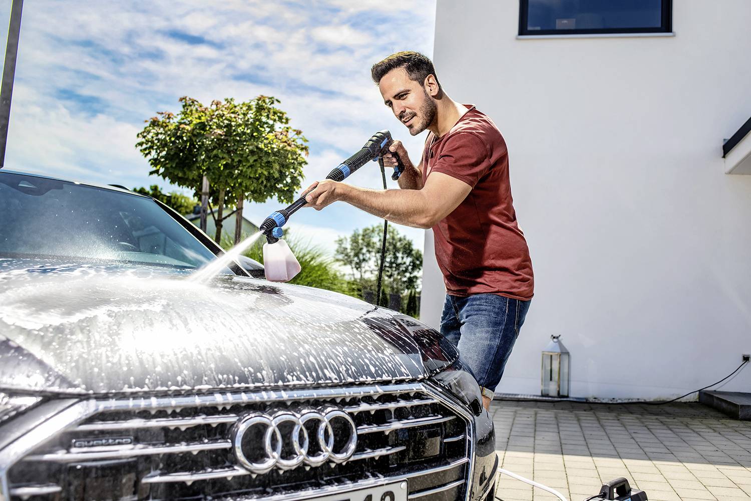 Muž umýva auto vysokotlakovým čističom na príjazdovej ceste. V pozadí je vidieť moderný dom.