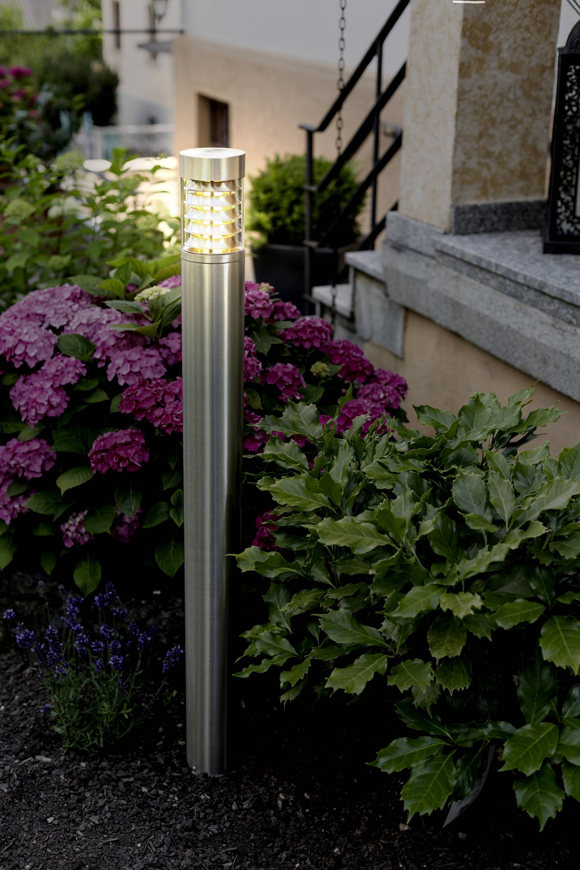 Moderná vonkajšia lampa stojí v záhrade, obklopená ružovými hortenziami a zelenými listami, vedľa kamenného schodiska.