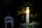 Kovová záhradná lampa svieti v noci vedľa starého dreveného púzdra lampy, obklopená tmavým lístím.
