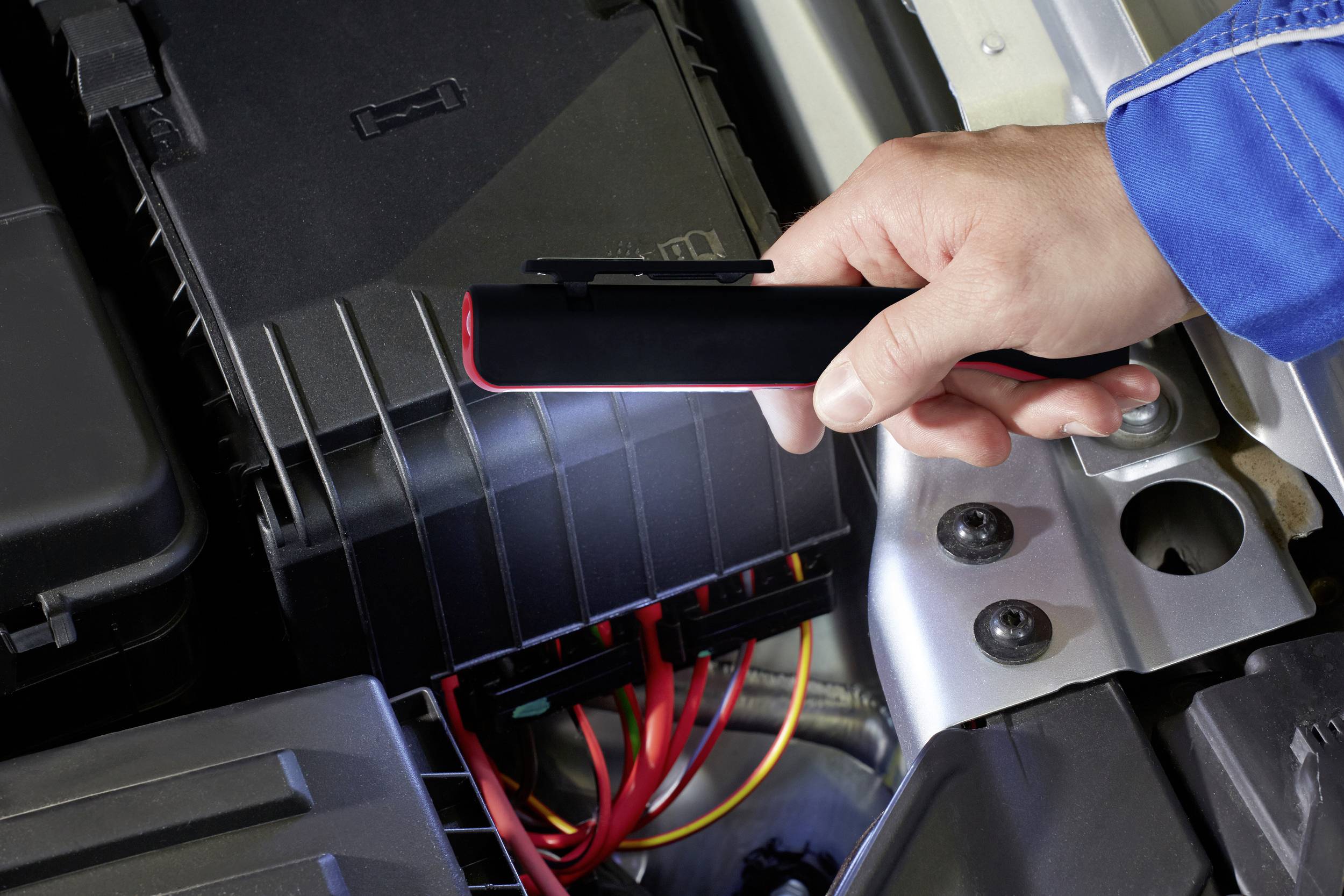 Osoba drží lampu a osvetľuje otvorený motorový priestor s viditelnými káblami a krytom batérie.