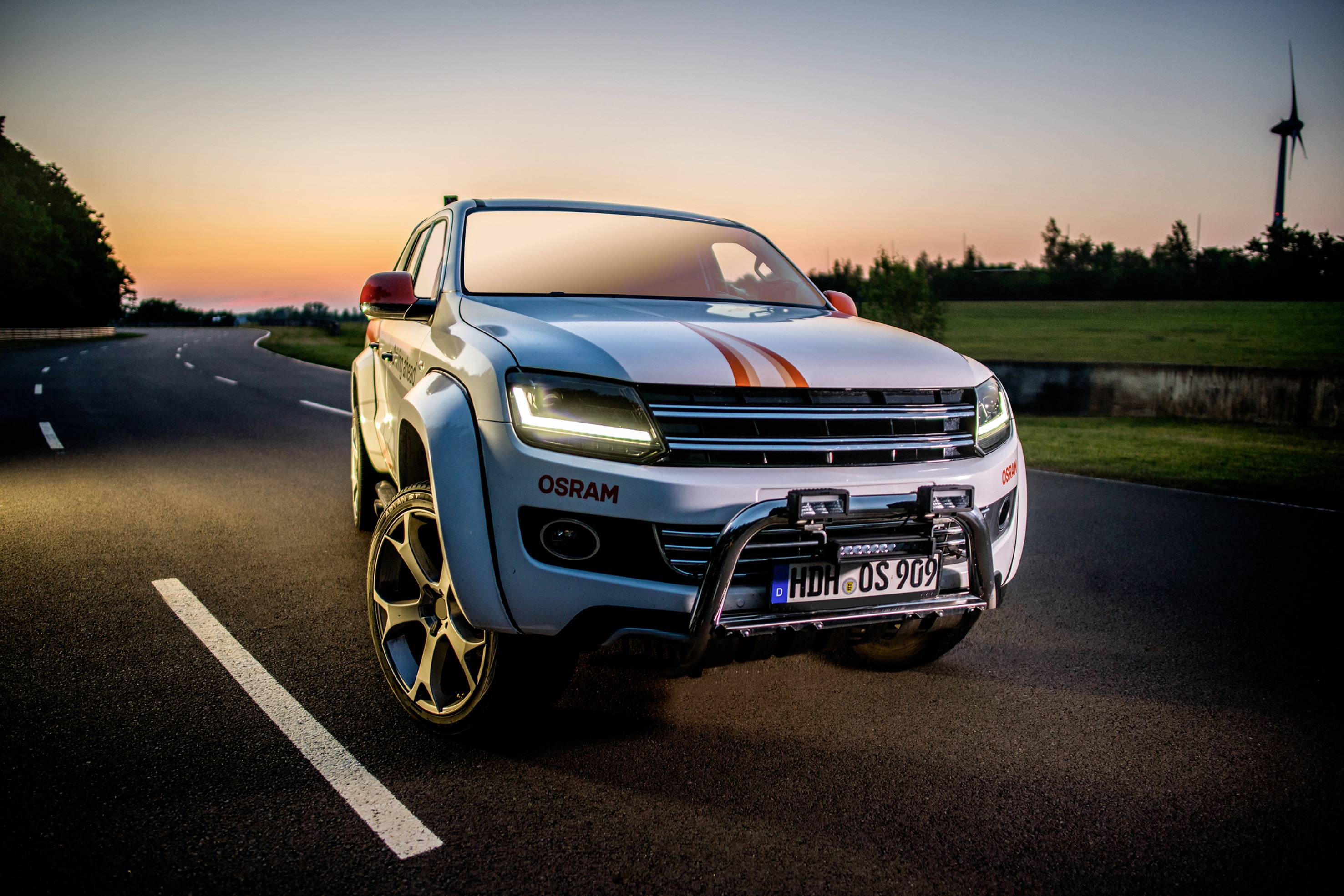 Biele SUV s oranžovými pruhmi stojí na prázdnej ceste počas západu slnka, v pozadí veterná turbína.