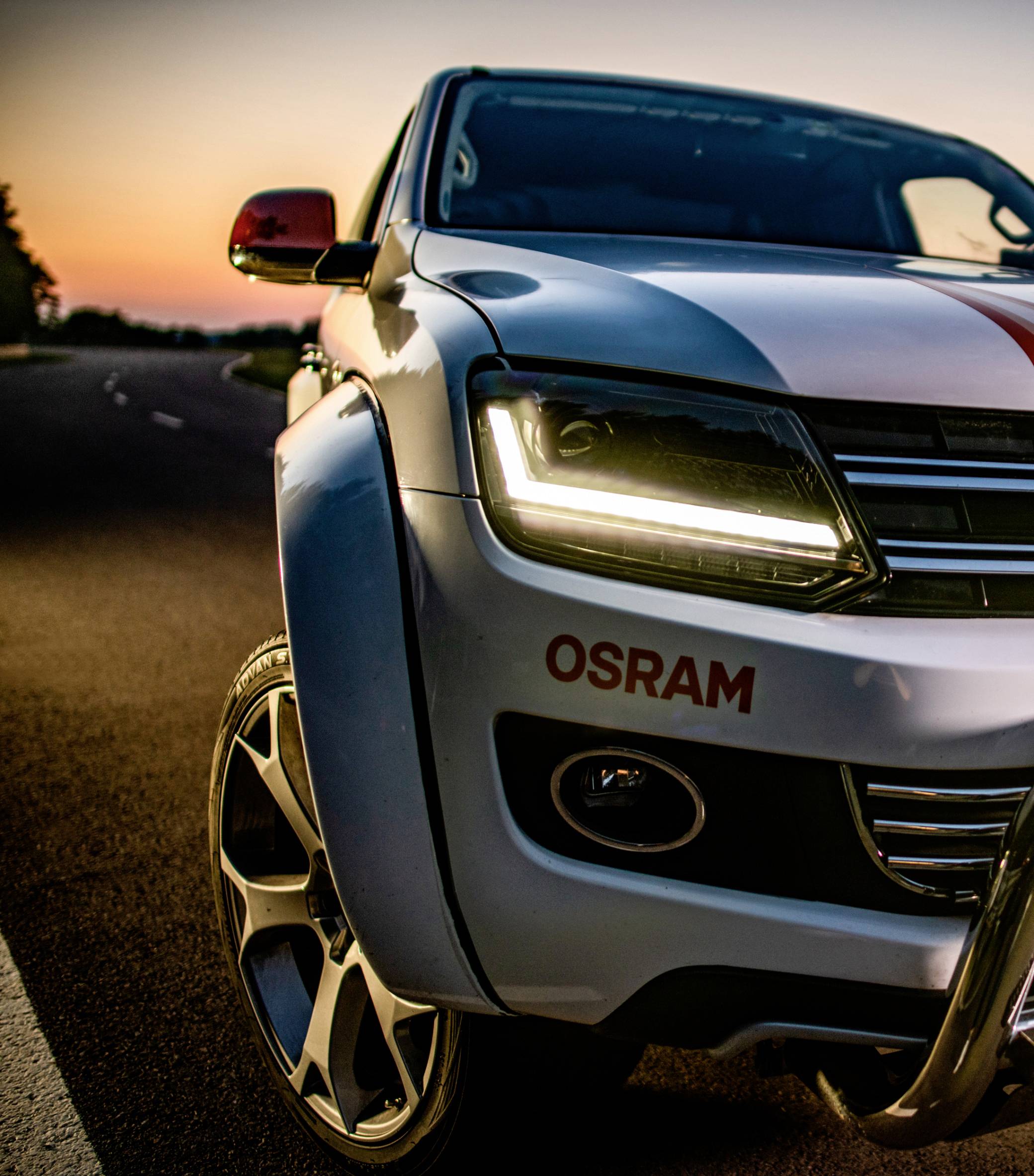 Biely pickup truck s logom OSRAM stojí na ceste pri západe slnka. Svetlomety sú zapnuté.