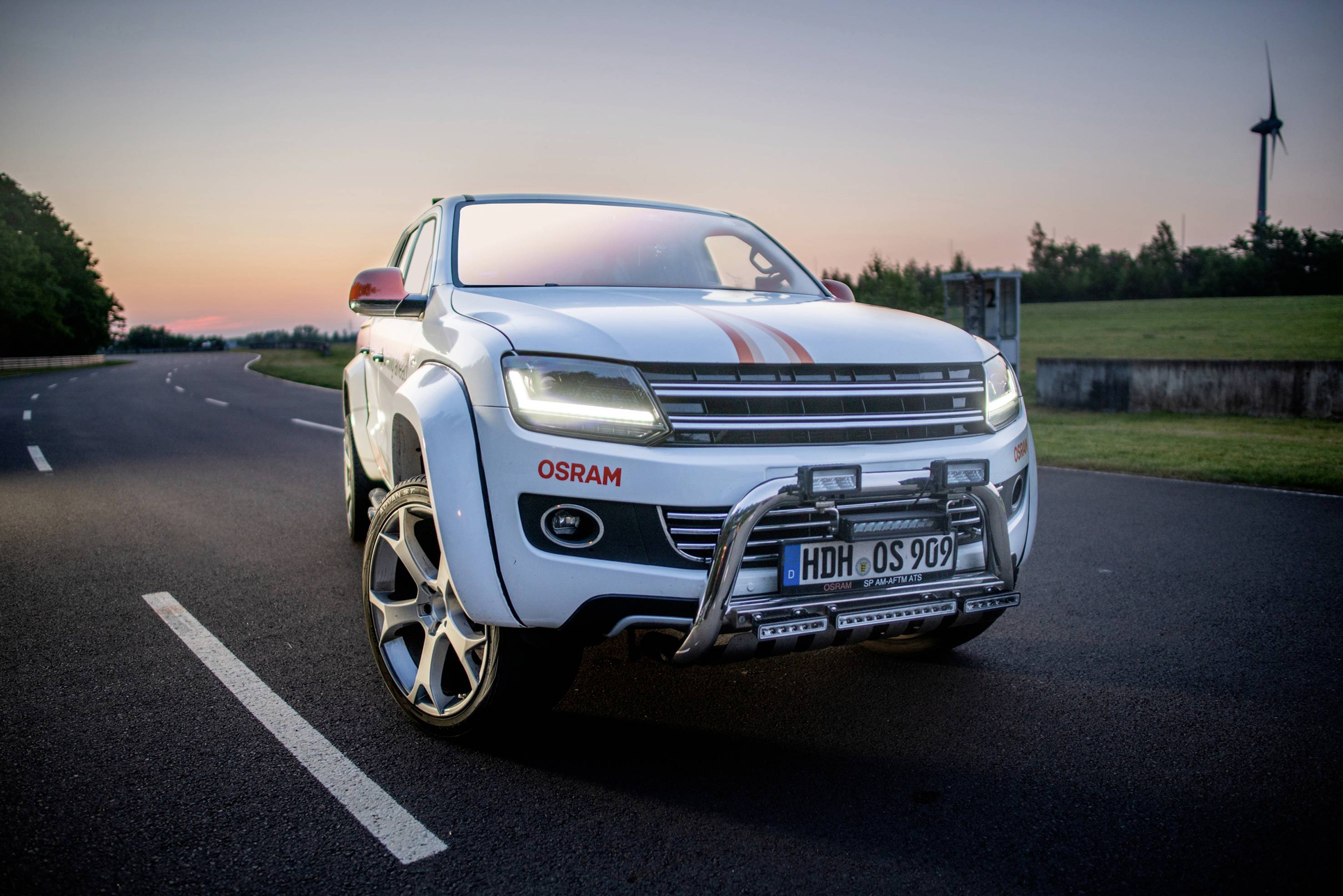 Biely SUV stojí na ceste pri západe slnka. Vozidlo má logo OSRAM, nápadné kolesá a bavorskú poznávaciu značku.