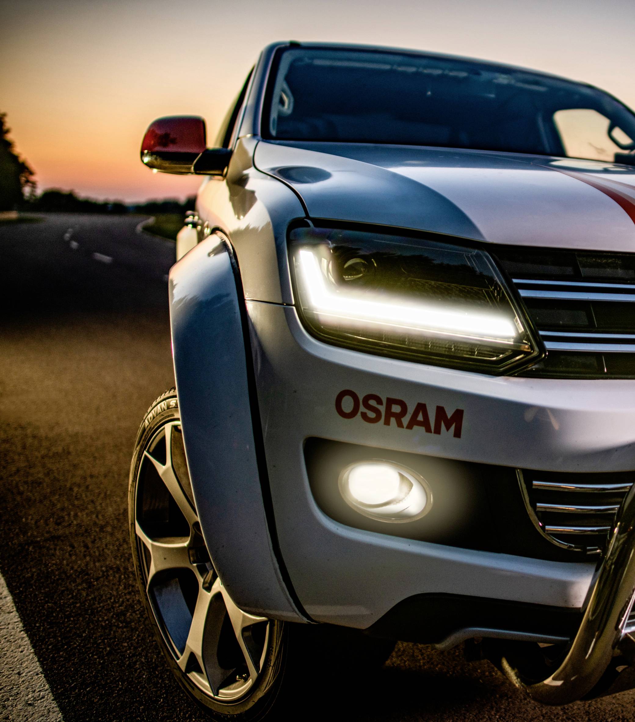 SUV v predvečerí na ceste, pohľad spredu so zapnutým svetlometom a nápisom 'OSRAM'.