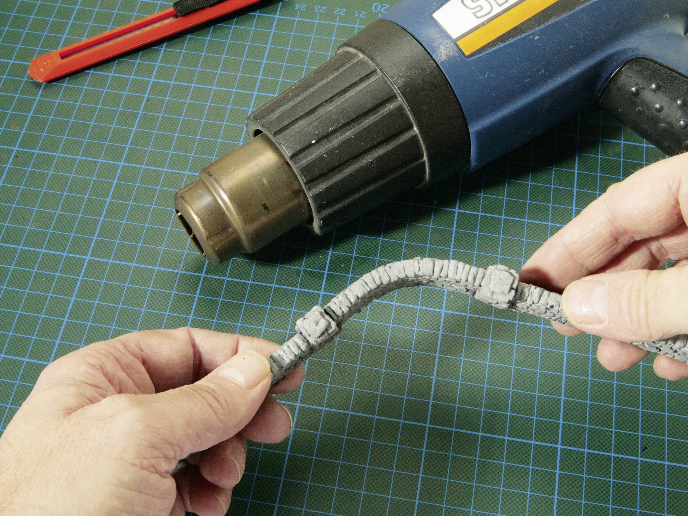 Osoba drží elektrický kábel, ktorý je opravovaný fénom na vlasy. V pozadí ležia pravítko a odlamovací nôž.