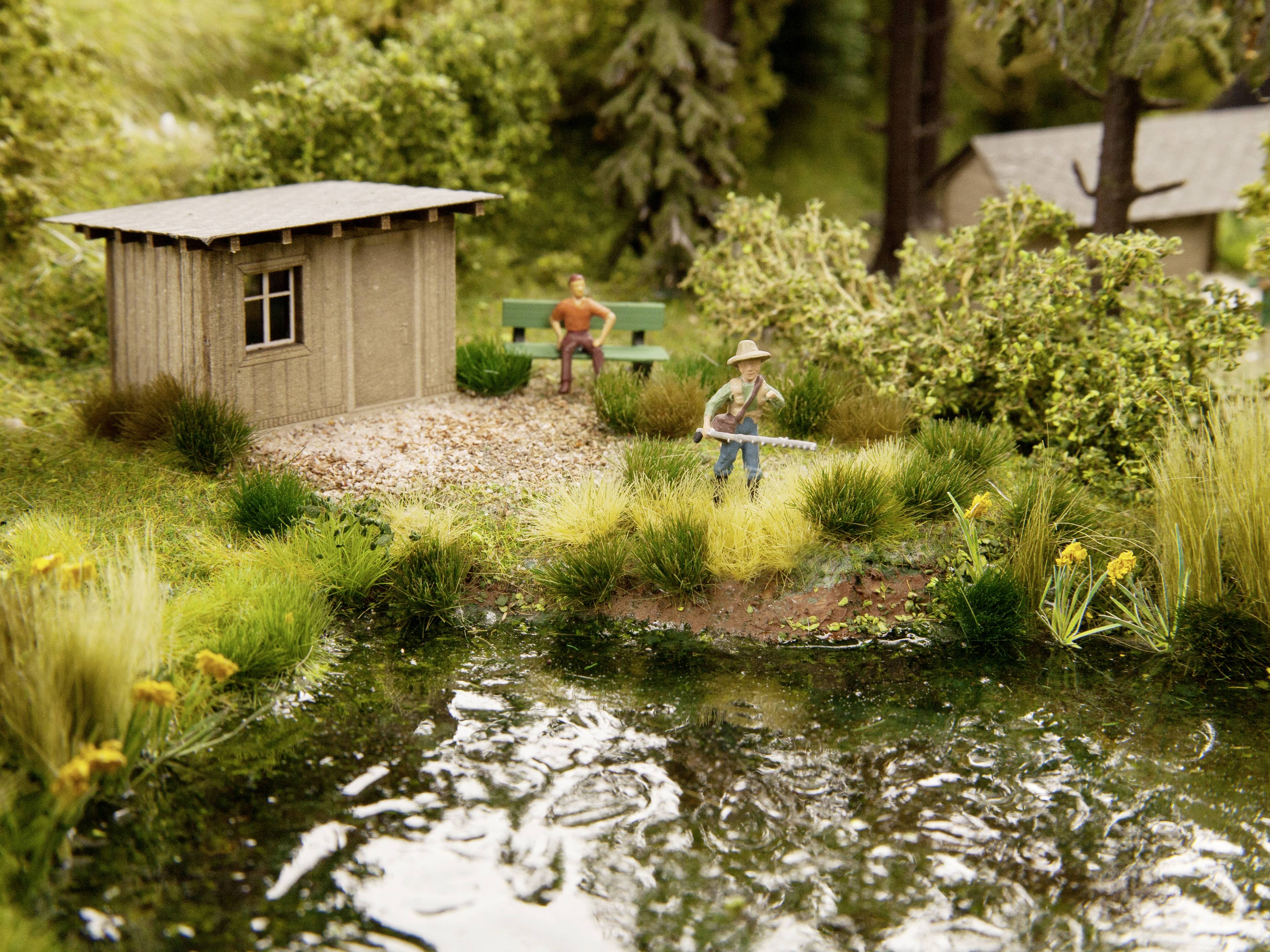 Miniaturový model zobrazuje vidiecku scénu pri rybníku, s rybárom v popredí a osobou sediacom na lavičke v pozadí.