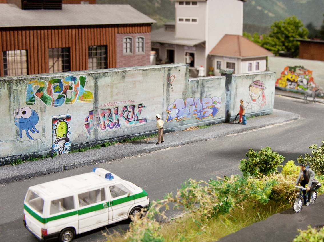 Pouličná scéna s pomaľovaným múrom, okoloidúcimi, policajným autom a bicyklami. Živé graffiti zdobí múr, obklopený budovami.