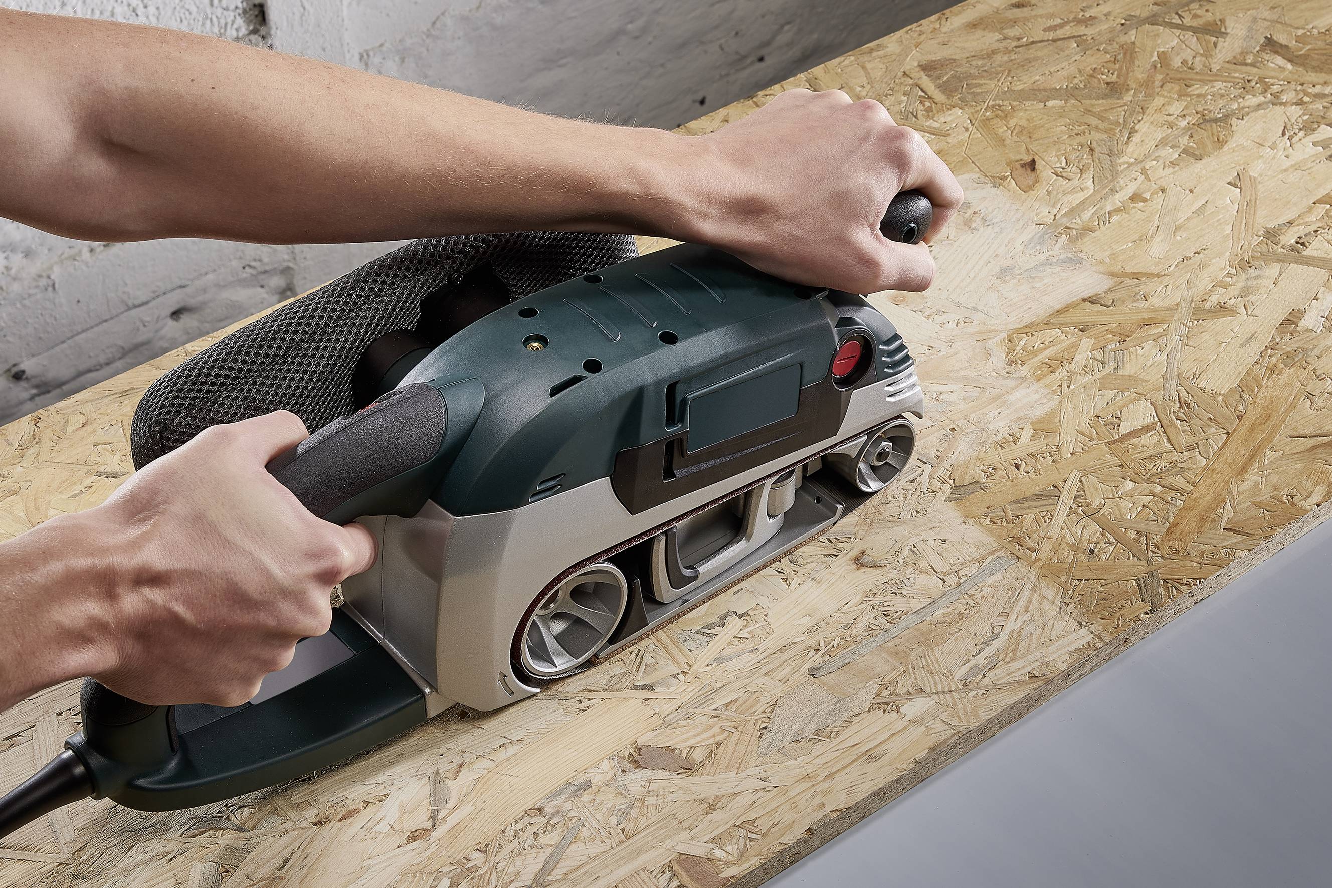 Osoba brúsi drevotrieskovú dosku elektrickým pásovým brúsnym strojom. Záber sa sústreďuje na ruky a proces brúsenia.