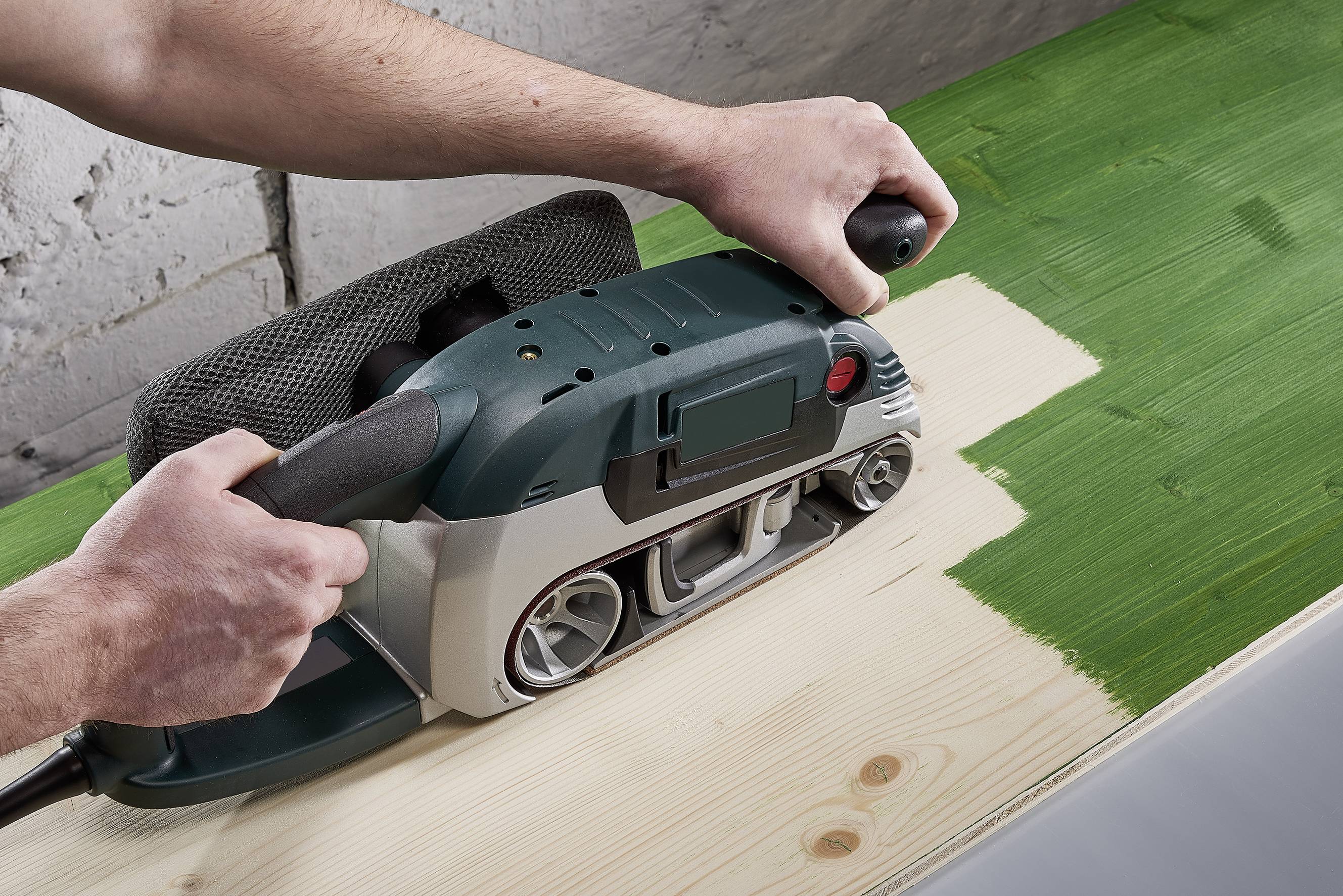 Osoba brúsi drevo pásovou brúskou. Povrch ukazuje zmenu farby z zelene na prírodné drevo.