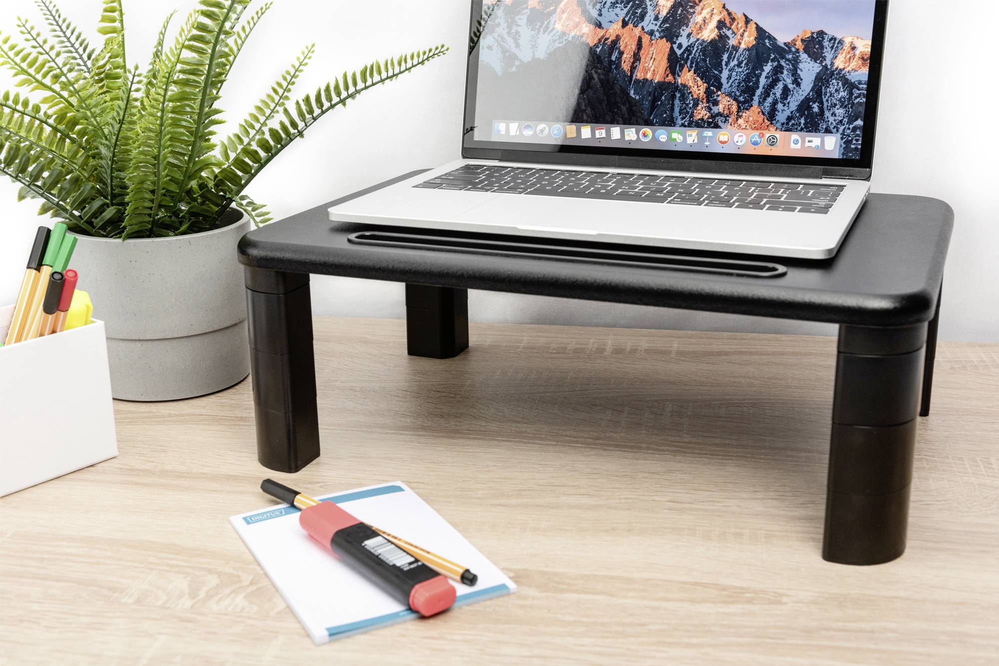 Laptop stojí na čiernom stolnom stojane. Vedľa stola sa nachádza kvetináč s rastlinou a kancelárske potreby.