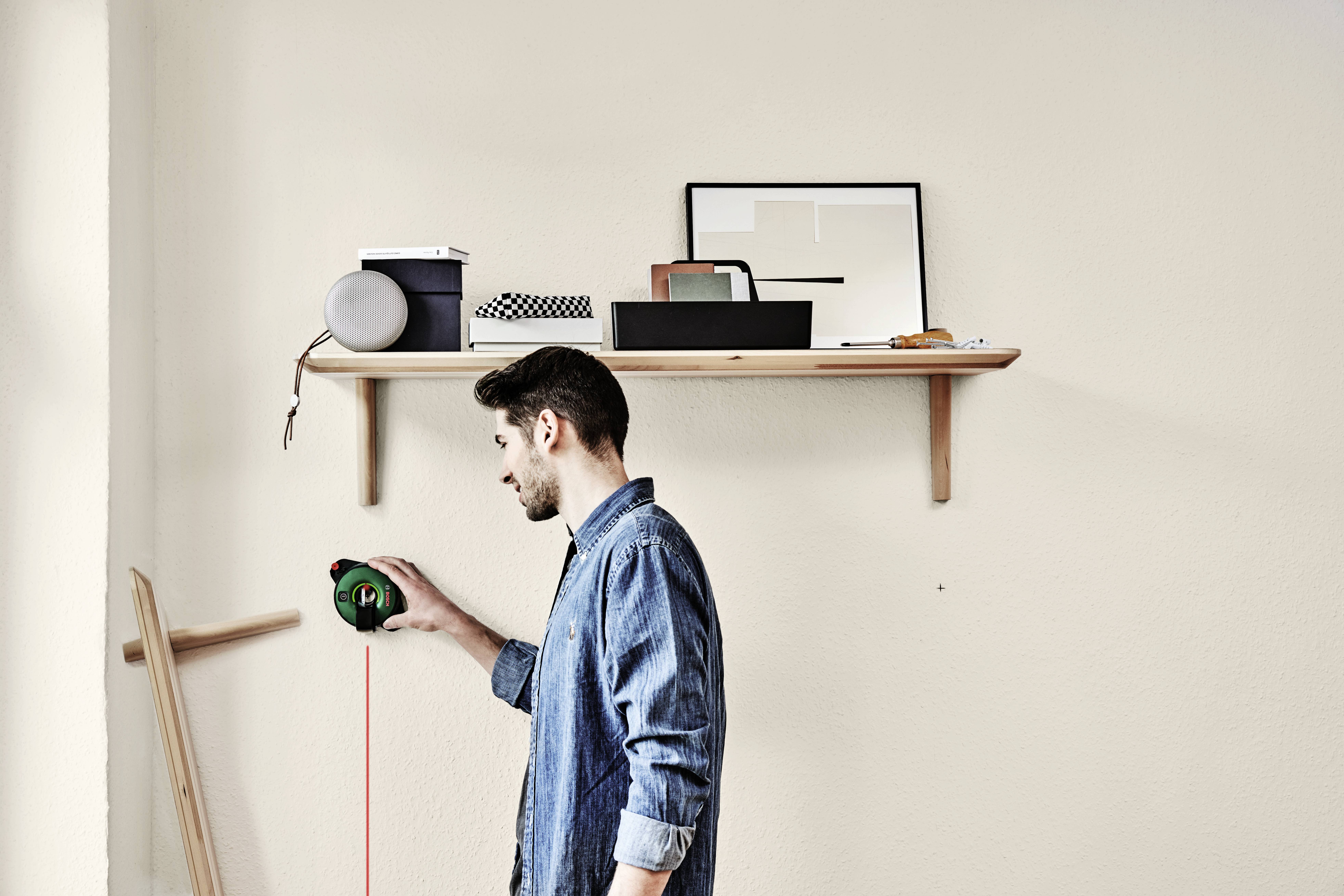 Muž meria výšku steny pomocou meracieho pásma. V pozadí visí polica s rôznymi predmetmi.