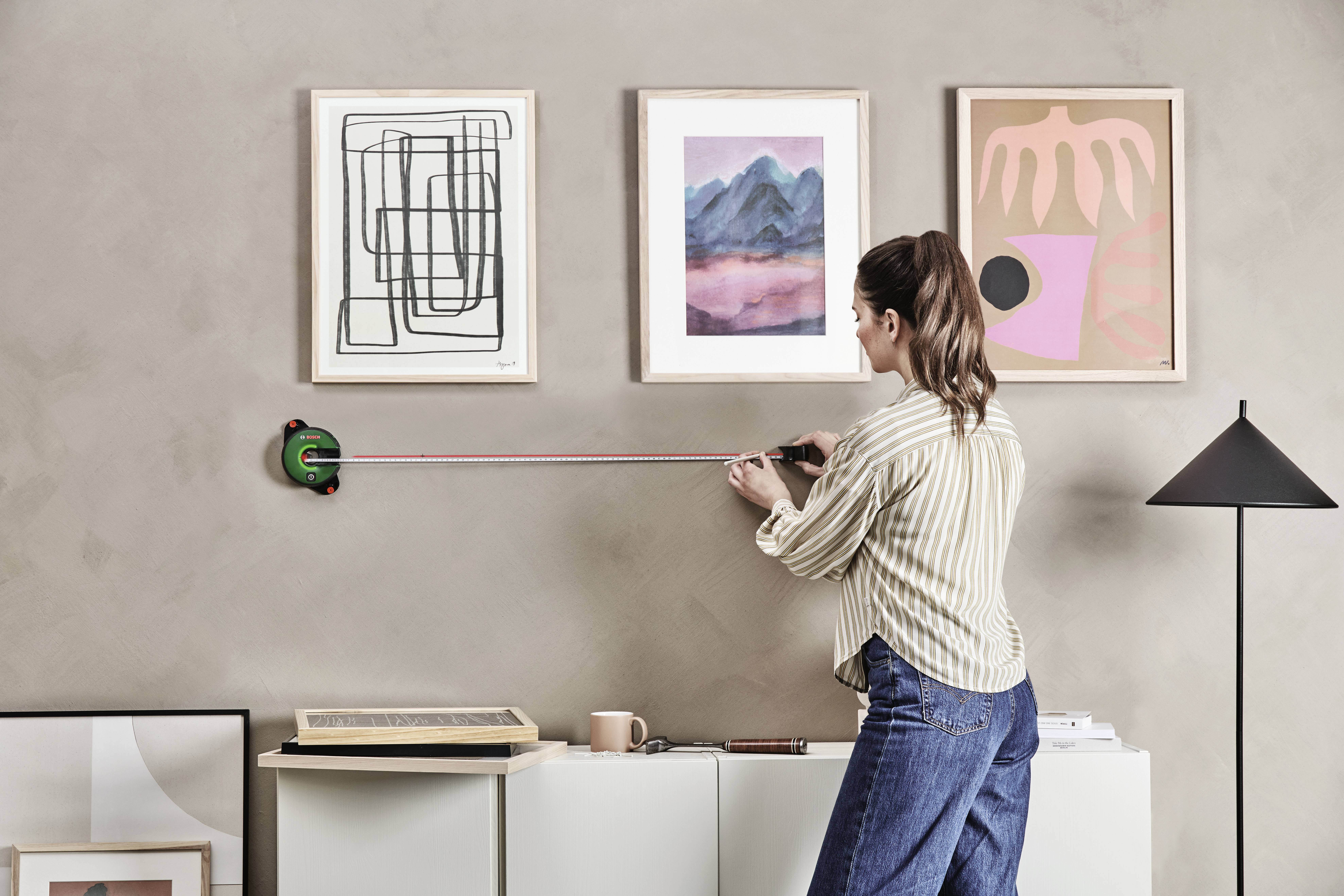 Žena meria výšku pre obrazy na stene pomocou meracieho pásma. Na stene visia tri zarámované umelecké diela, vrátane abstraktných a krajinárskych diel.