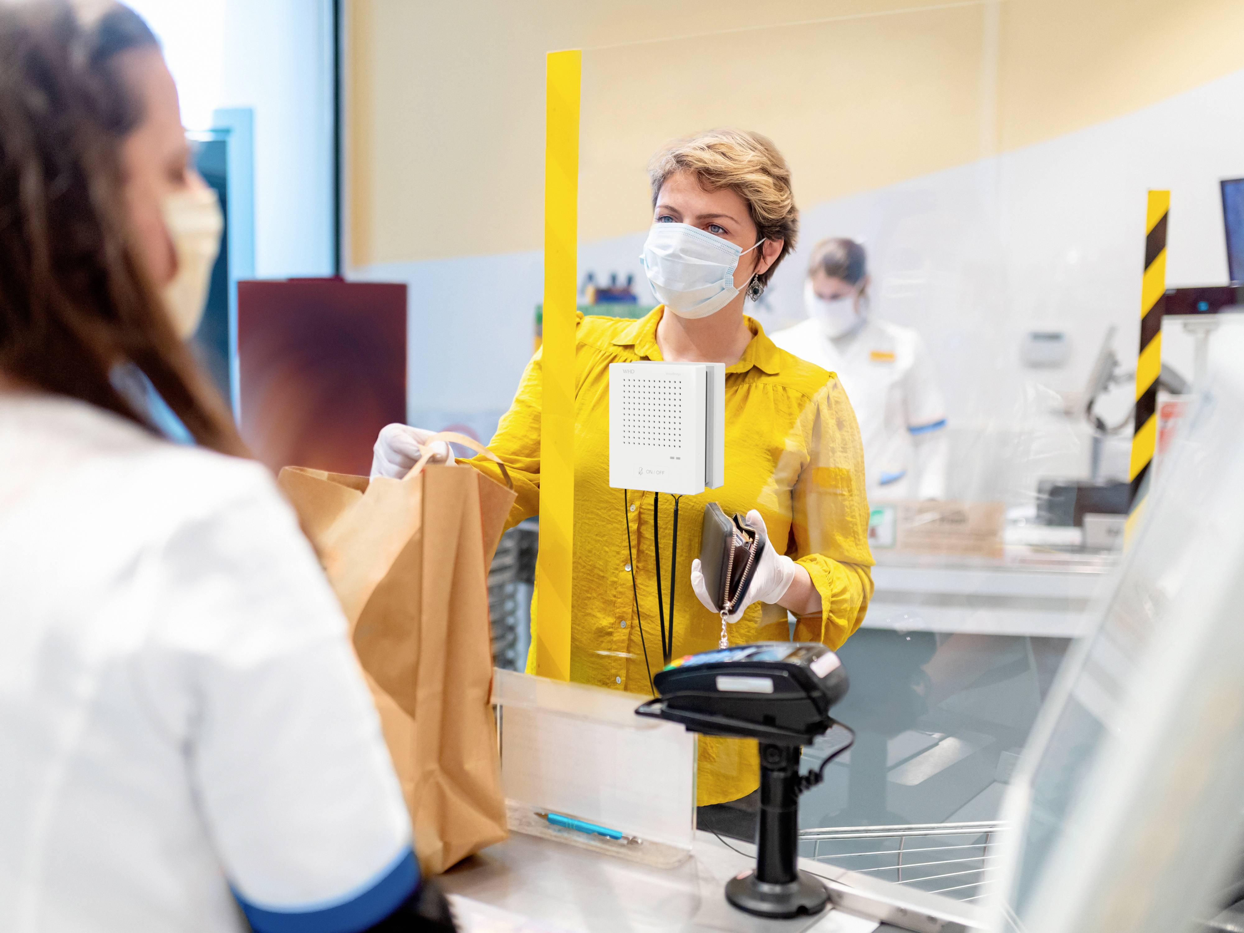 Osoba v žltom oblečení obsluhuje pokladňu v supermarkete, zatiaľ čo iná osoba s rúškom čaká. Medzi nimi je inštalované plexisklové oddeľovacie sklo.